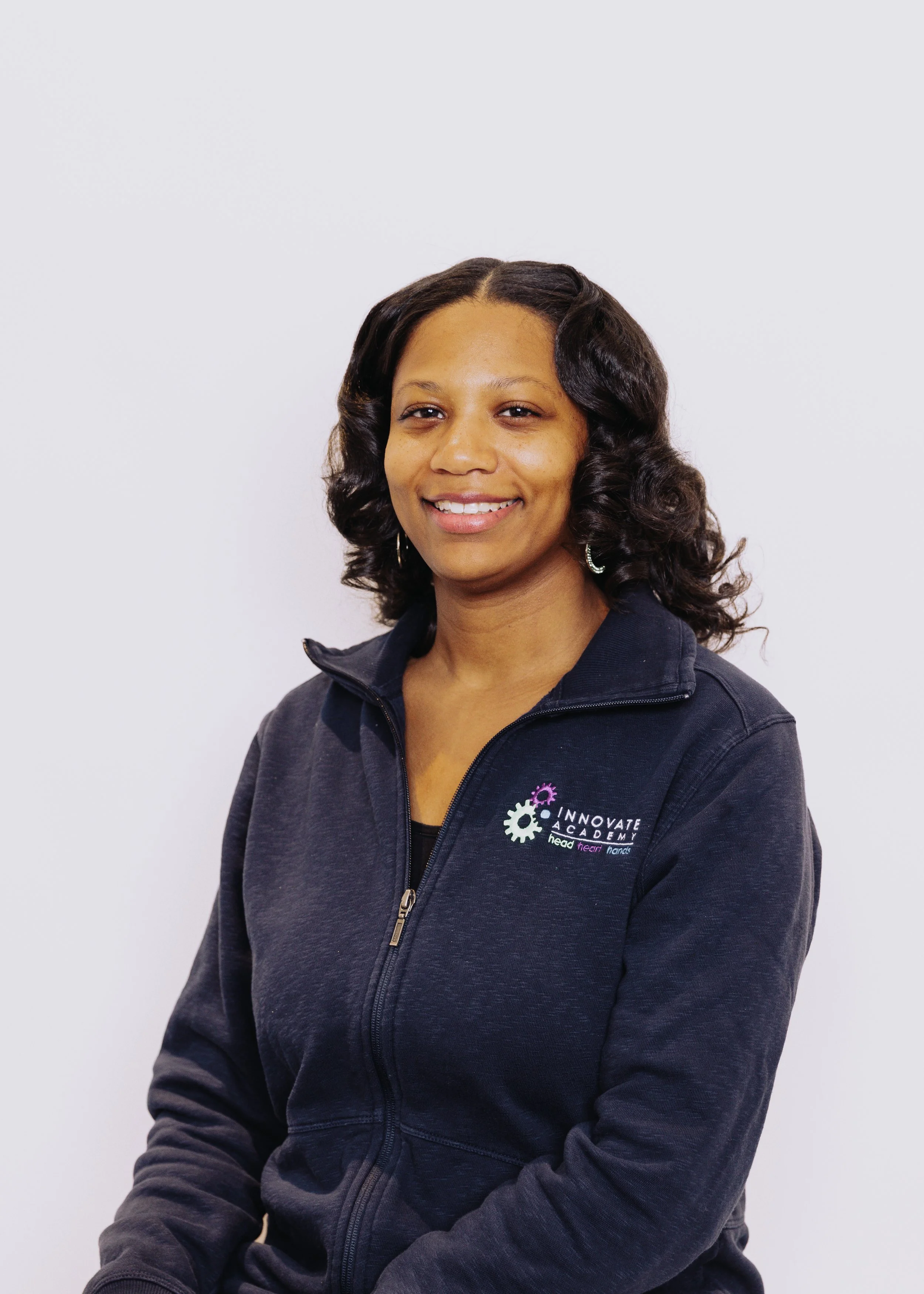 A woman with shoulder-length curly hair, smiling, wearing a black jacket with a logo that reads 'Innovate Academy' on the left chest, against a plain white background.