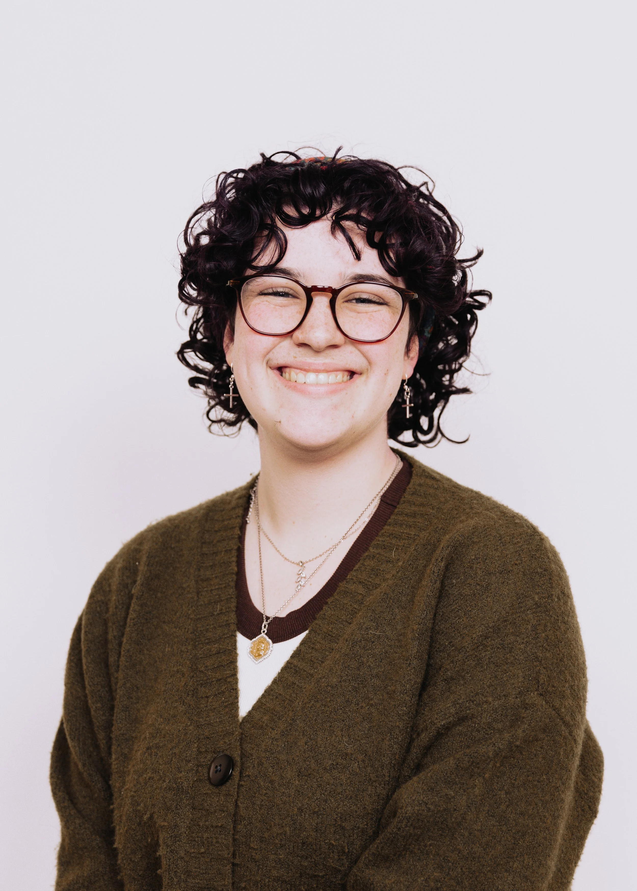 A young woman with curly dark hair, glasses, and earrings, smiling at the camera against a plain white background.