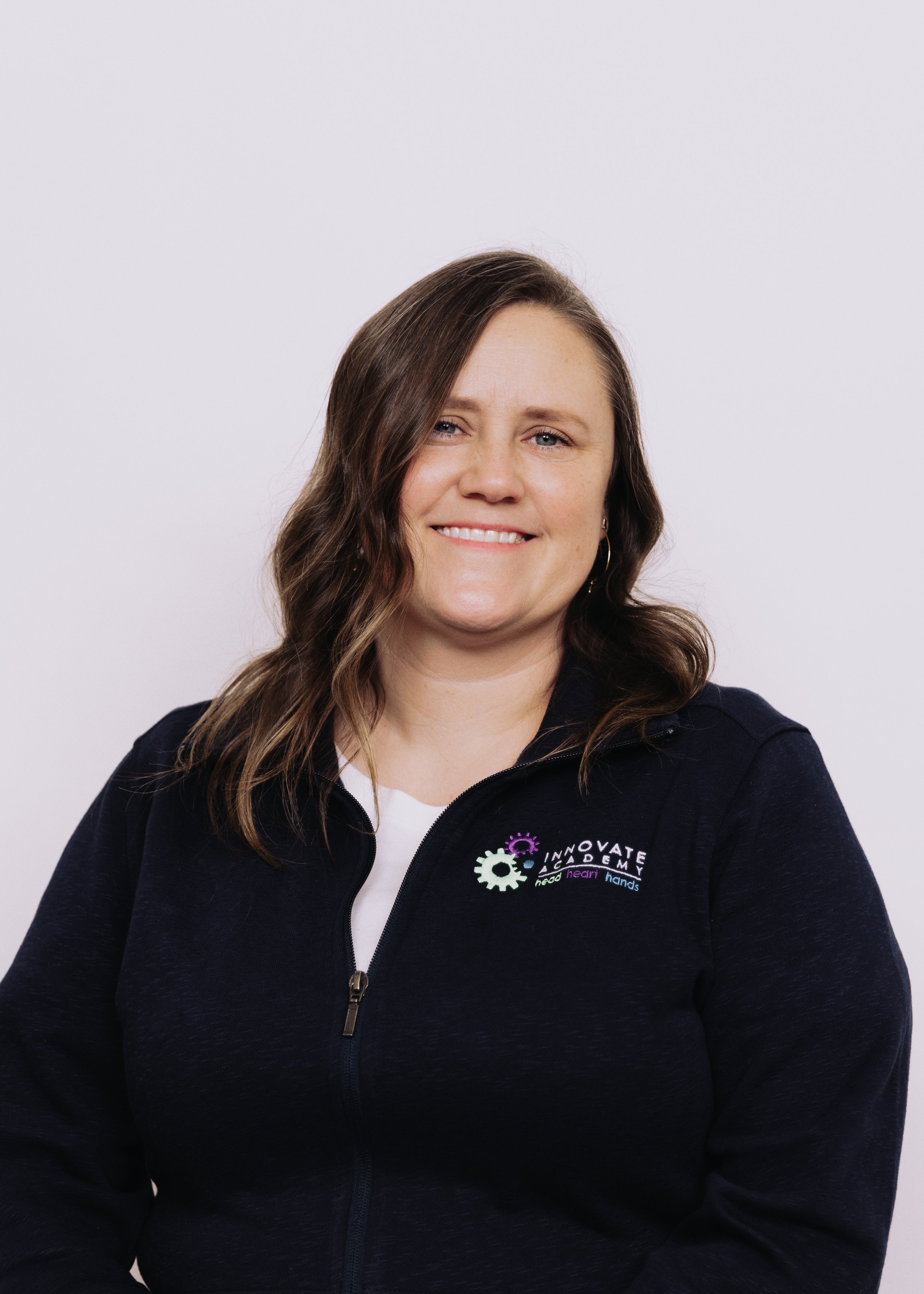 A woman with brown hair styled in waves, smiling, wearing a navy blue zip-up jacket with a logo that says "Innovate Academy" and the words "head, learn, hands" underneath, standing against a plain white background.