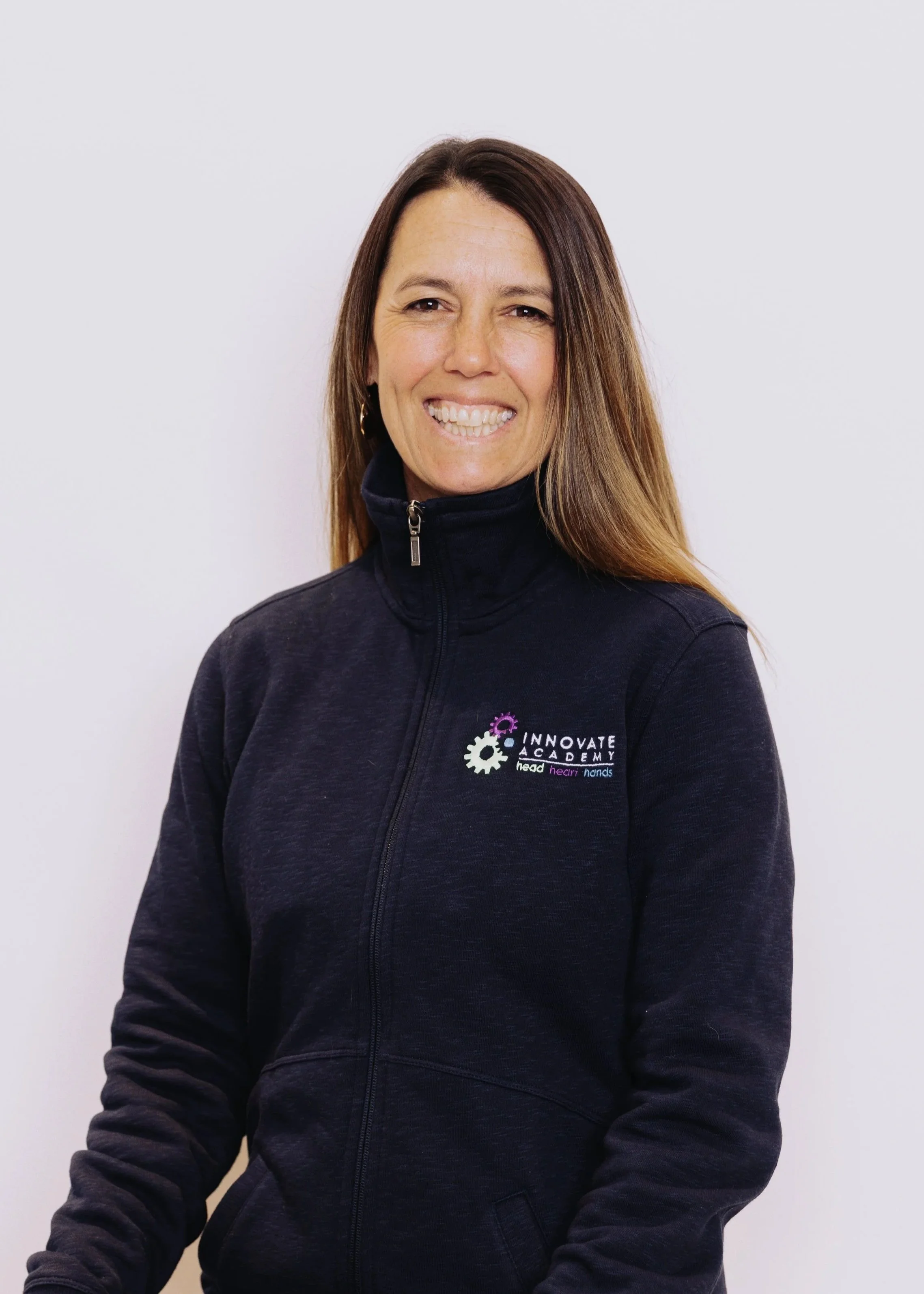 A woman smiling, wearing a black zip-up jacket with a logo on the chest that says 'Innovate Academy' and includes the words 'head, heart, hands'. She has long brown hair and stands against a plain white background.