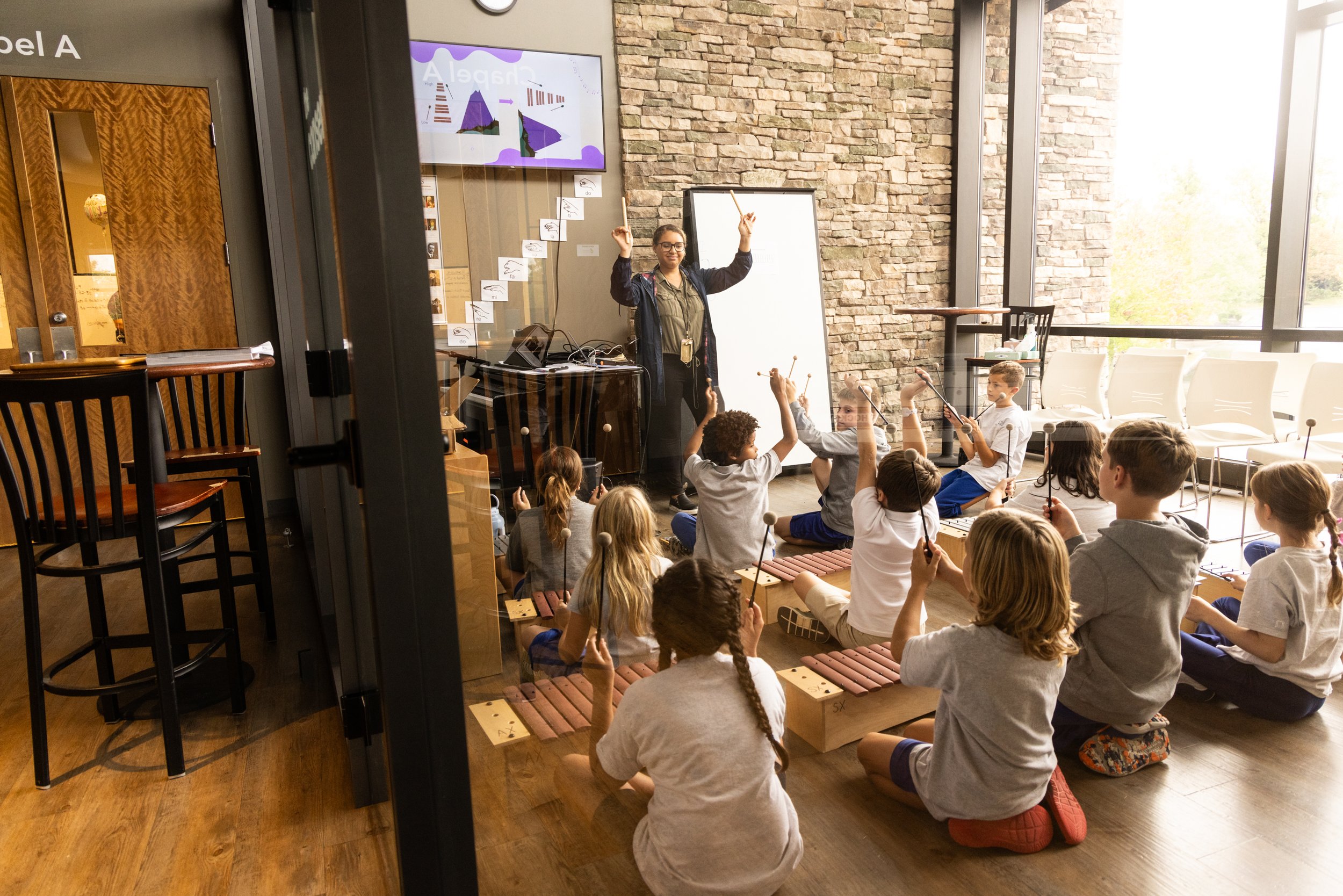 Children participating in a music class, playing percussion instruments with a teacher in a room with brick walls and large windows.
