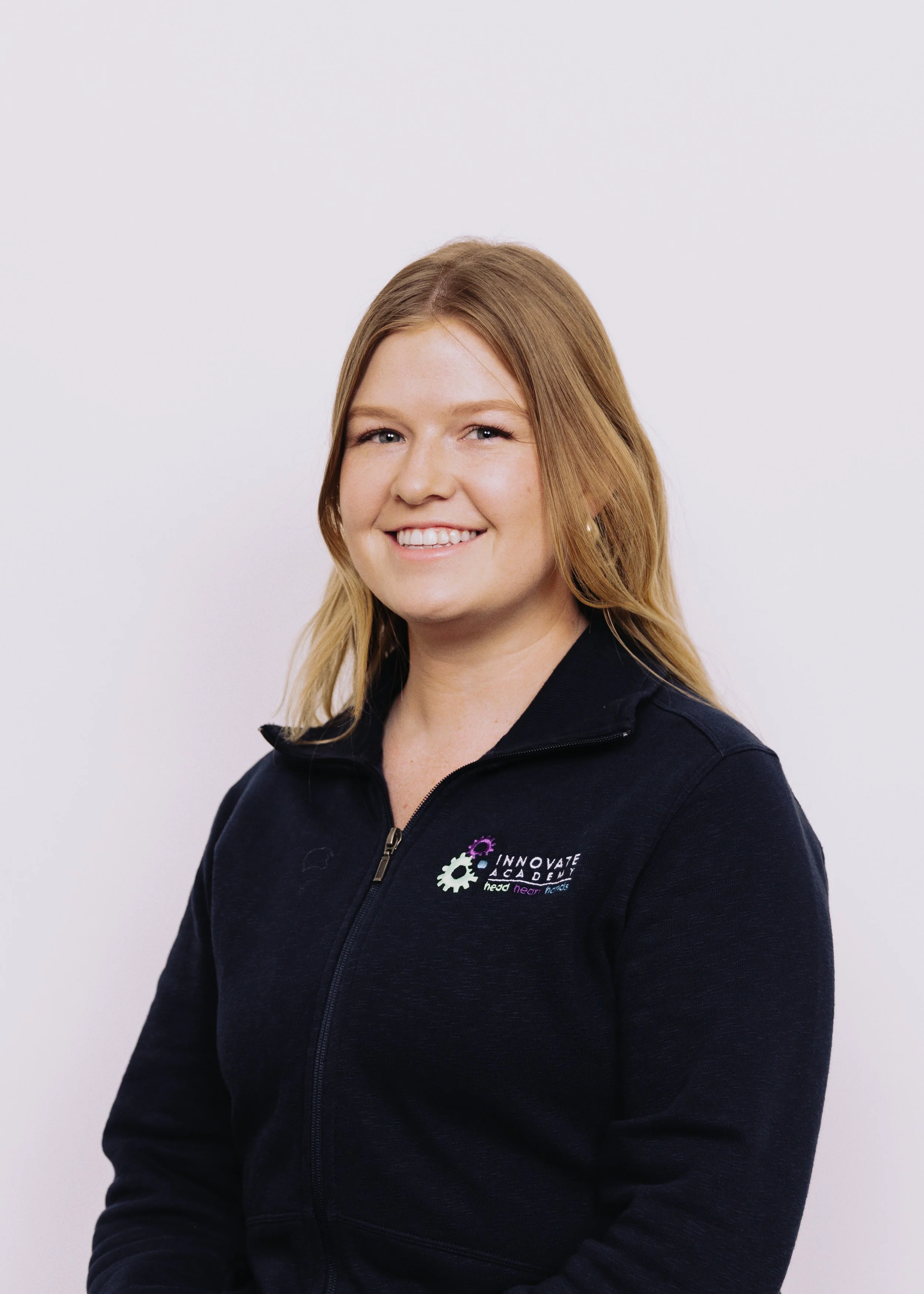 A young woman with shoulder-length blonde hair smiling, wearing a black zip-up jacket with a logo that reads "INNOVATE ACADEMY" on the left chest area, standing against a plain white background.