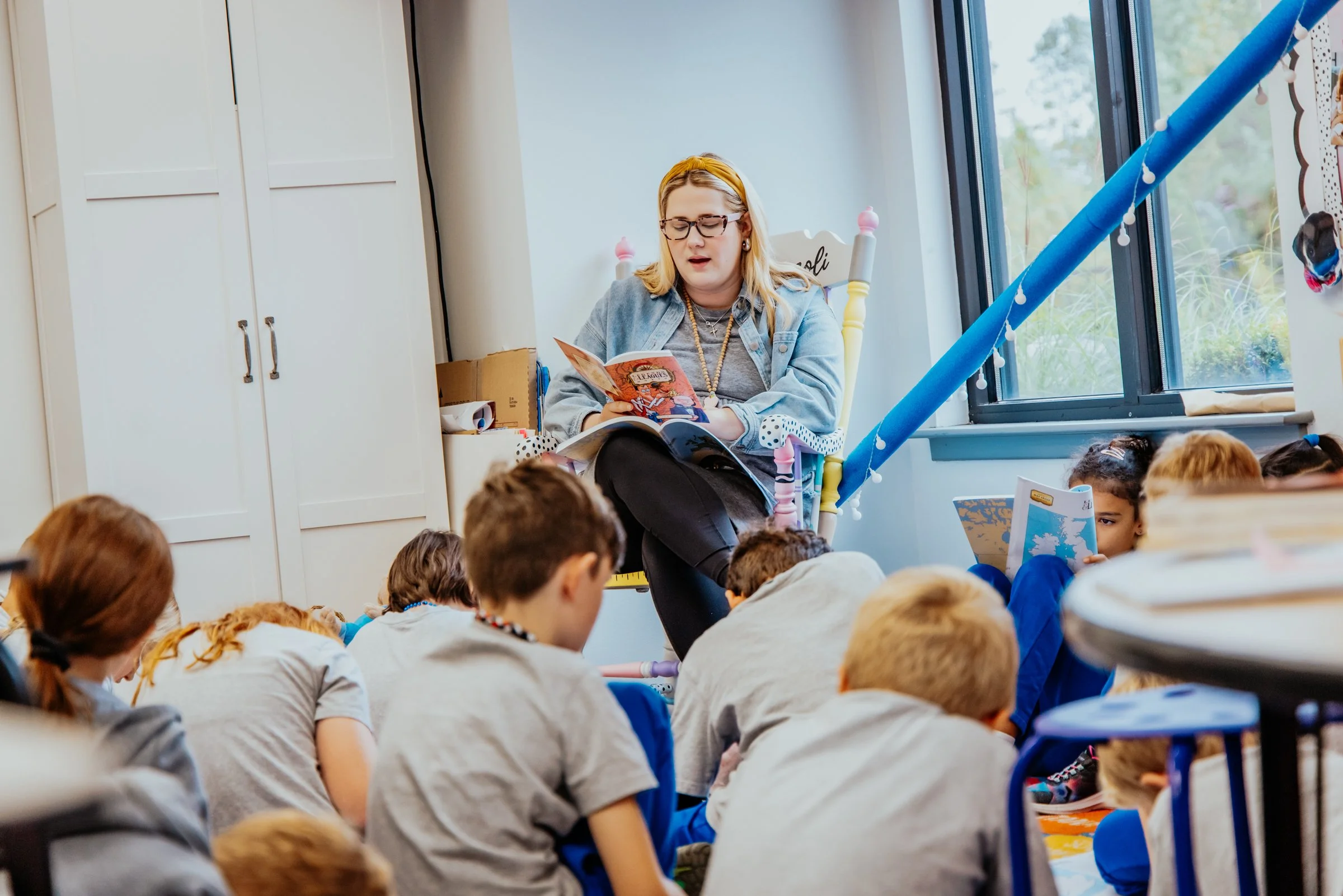 A woman reading a book to a group of children in a classroom, sitting on the floor with the children attentively listening.