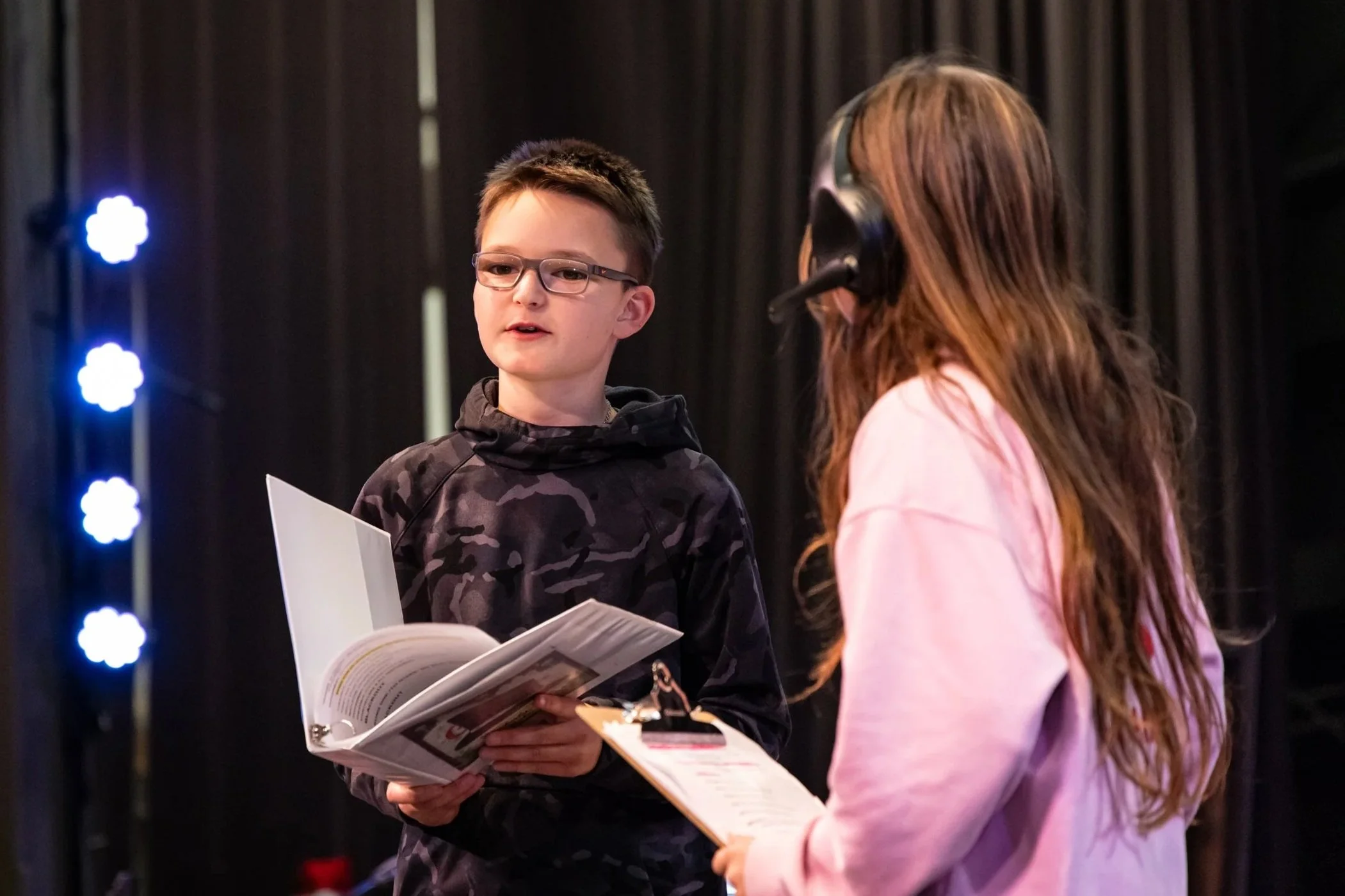 A young boy with glasses holding a script or booklet, talking to a girl wearing a headset, in a setting with stage lights and black curtains.