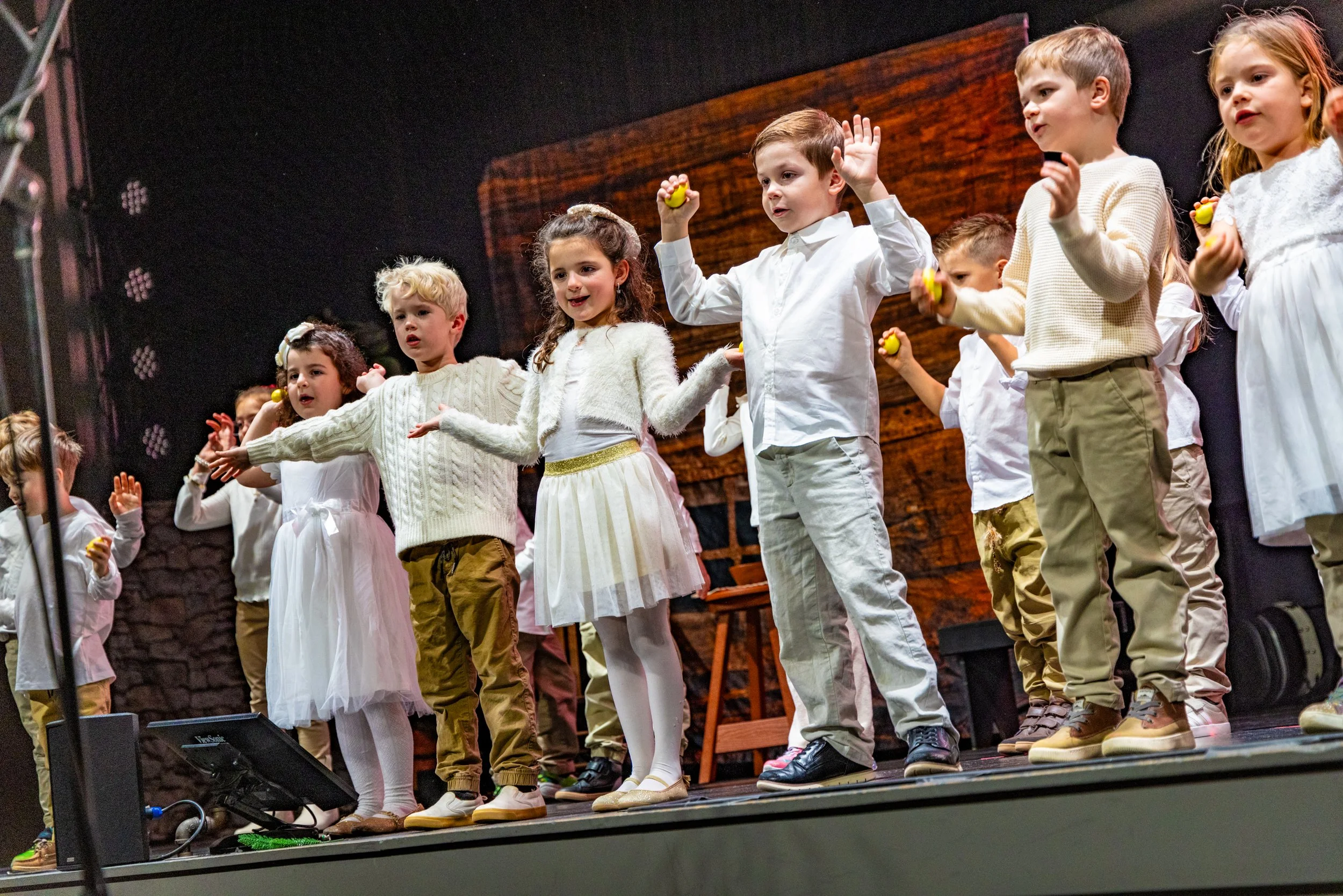 A group of children dressed in white and beige clothing standing on a stage, holding yellow objects, and participating in a performance or presentation.