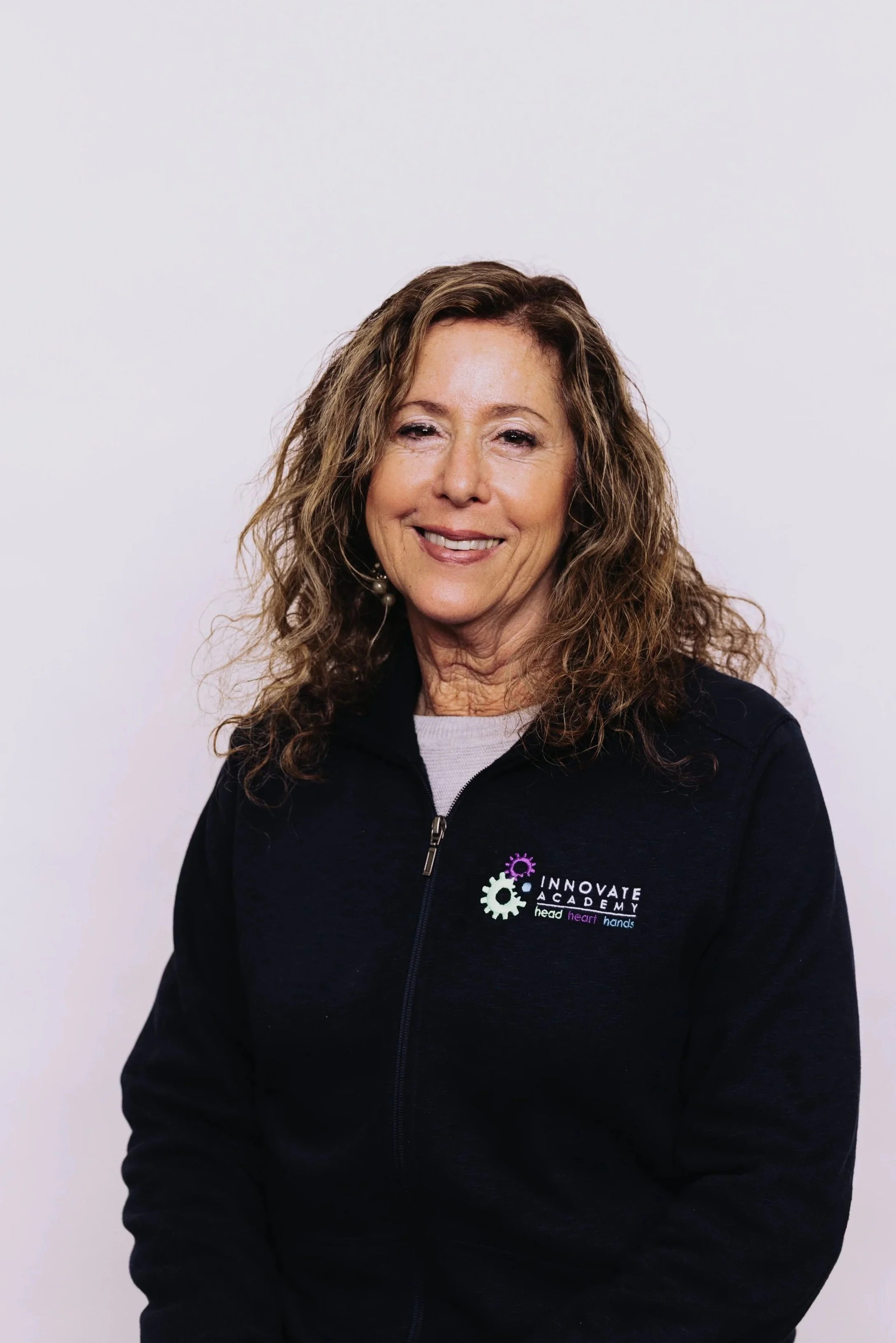 A woman with curly, reddish-brown hair smiling, wearing a black zip-up jacket with the "Innovate Academy" logo on it, against a plain white background.