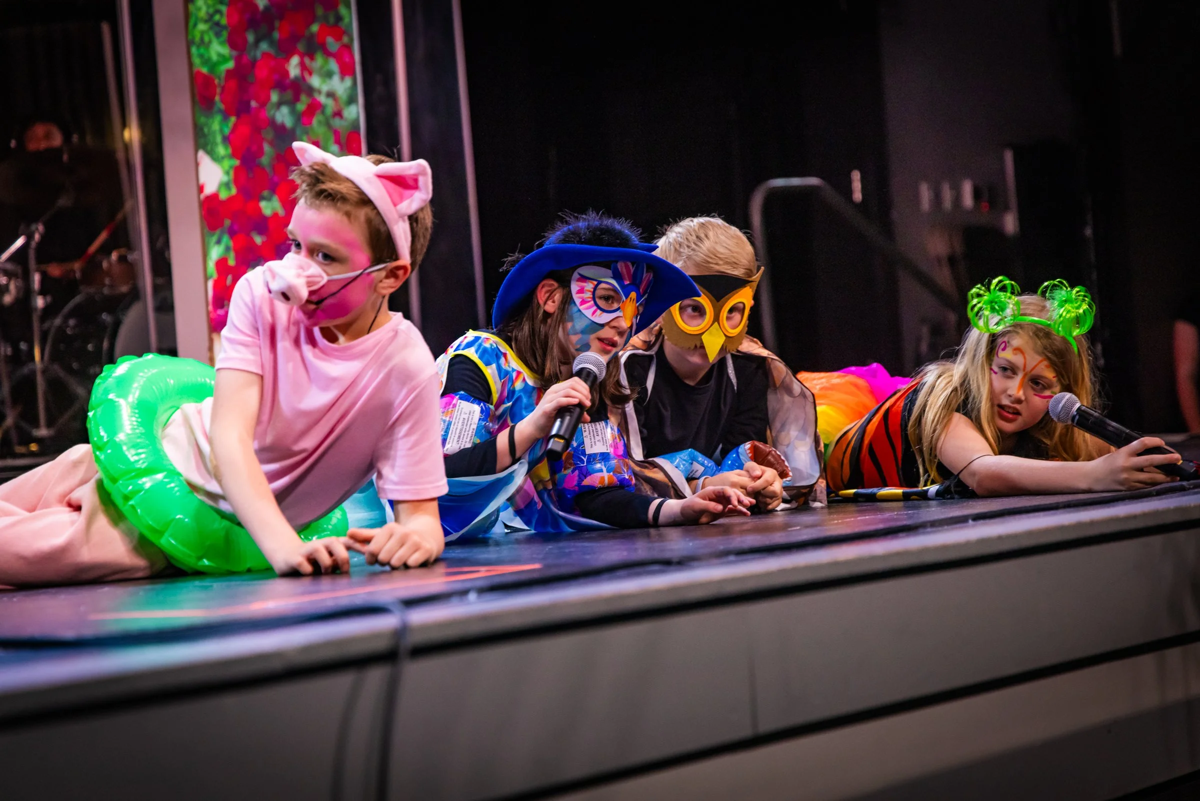 Children dressed in colorful costumes lying on stage, some wearing animal masks and accessories, participating in a performance or school event.