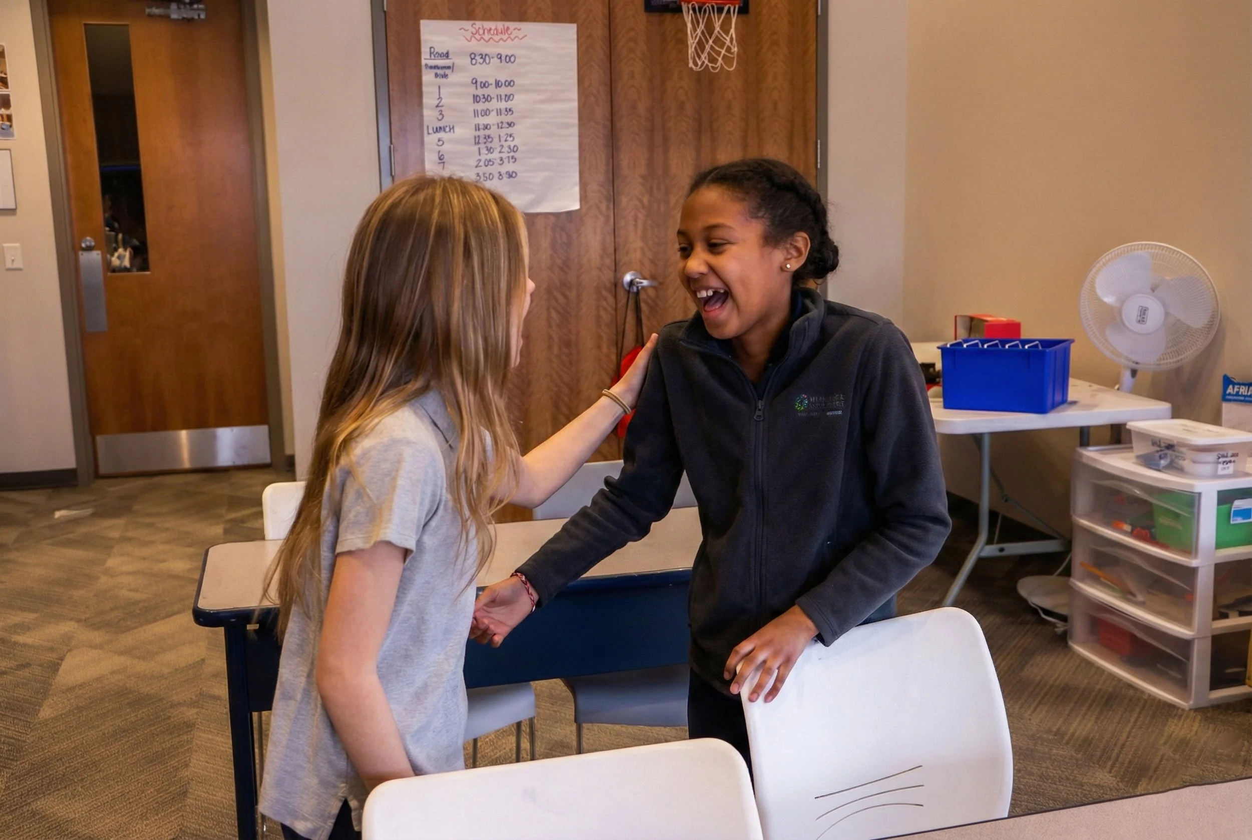 Two young girls are smiling and holding hands in a classroom. One girl is touching the other's shoulder with a lively expression. The classroom has a schedule posted on the door, a fan, and storage containers in the background.
