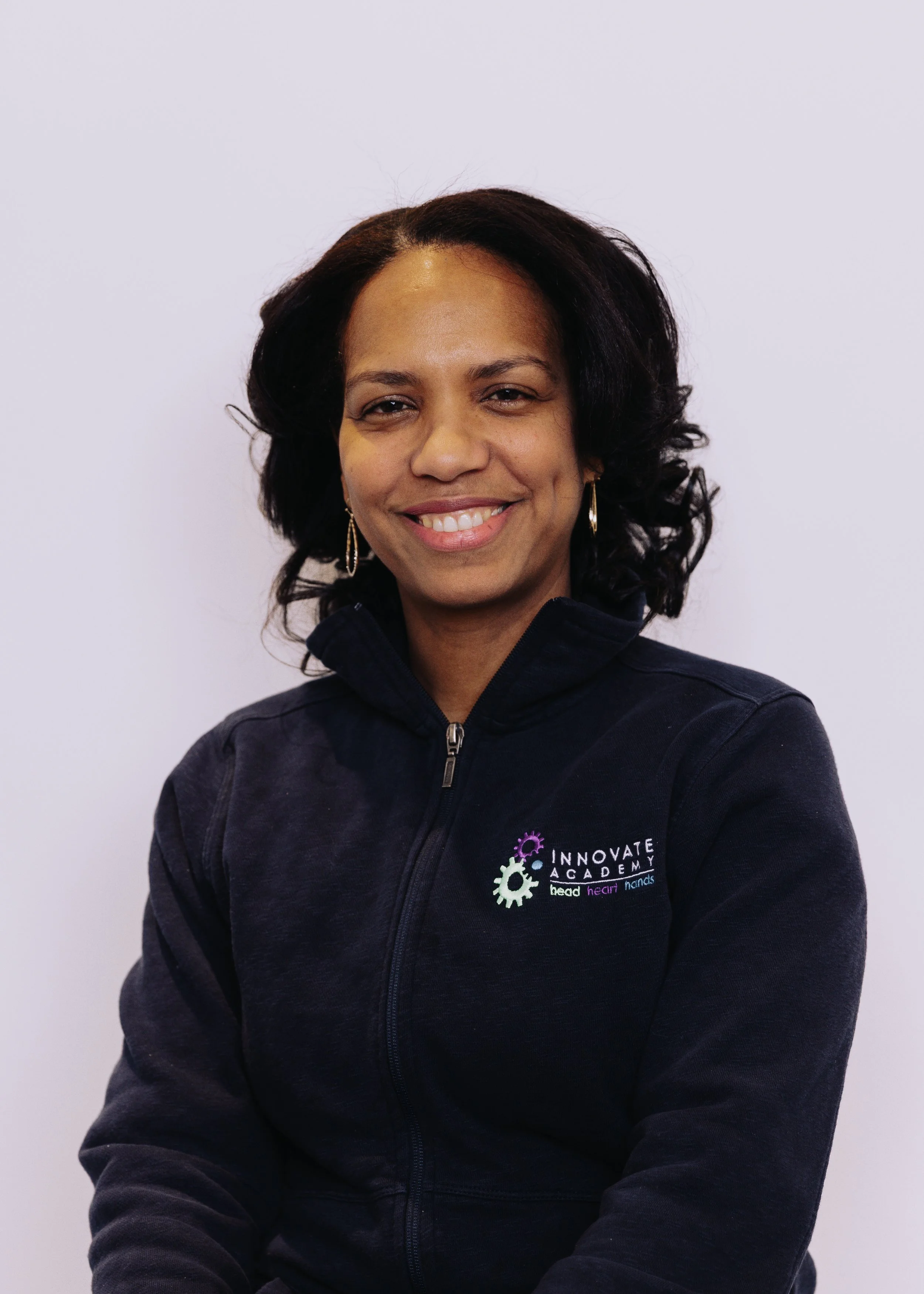 A smiling woman with black hair wearing a navy blue zip-up jacket with 'Innovate Academy' logo on the chest, standing against a plain white background.