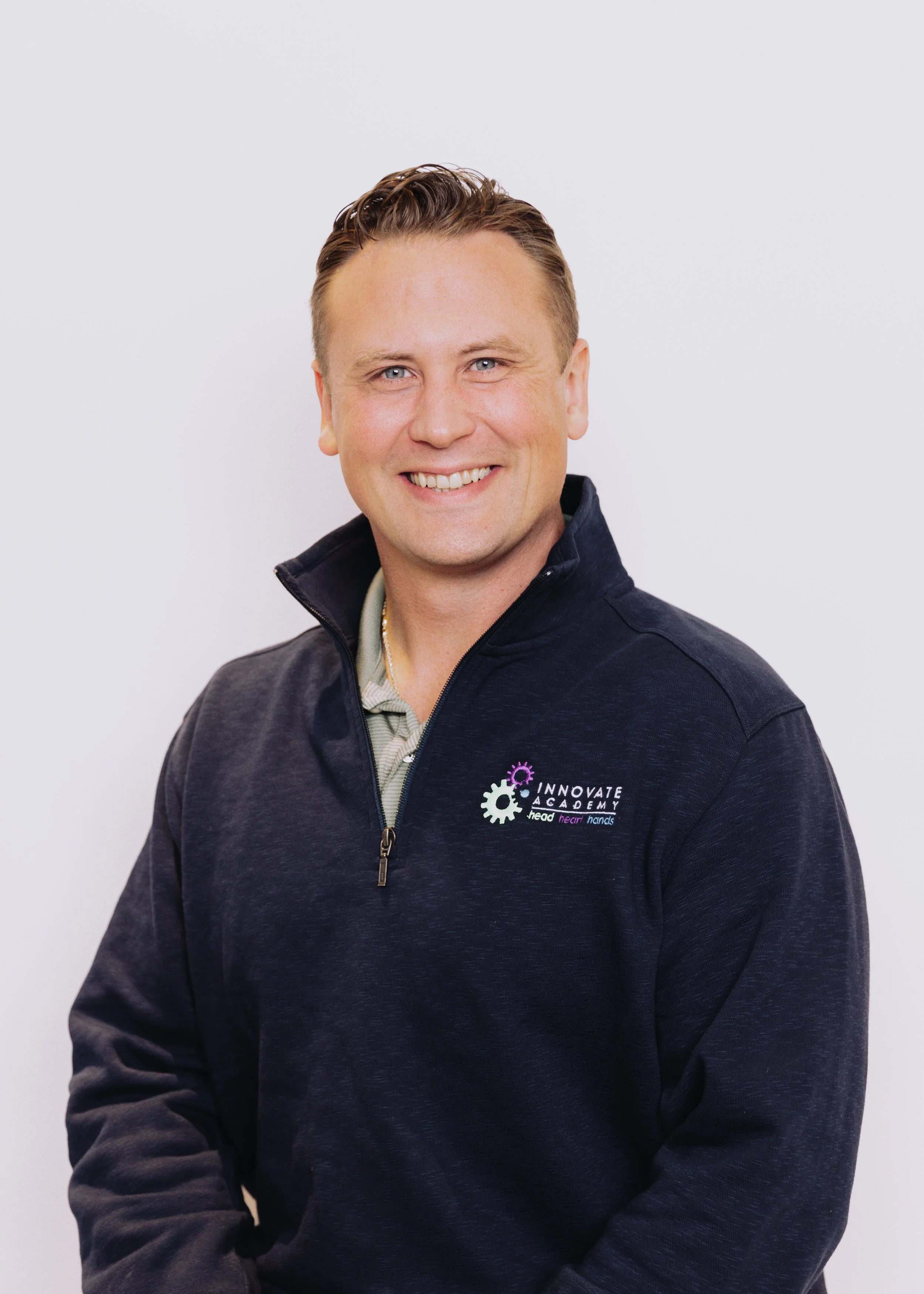 A smiling man wearing a dark navy zip-up jacket with an embroidered logo reading 'INNOVATE ACADEMY' and the tagline 'lead, hear, Hands'. He has short, styled blond hair and is standing against a plain white background.