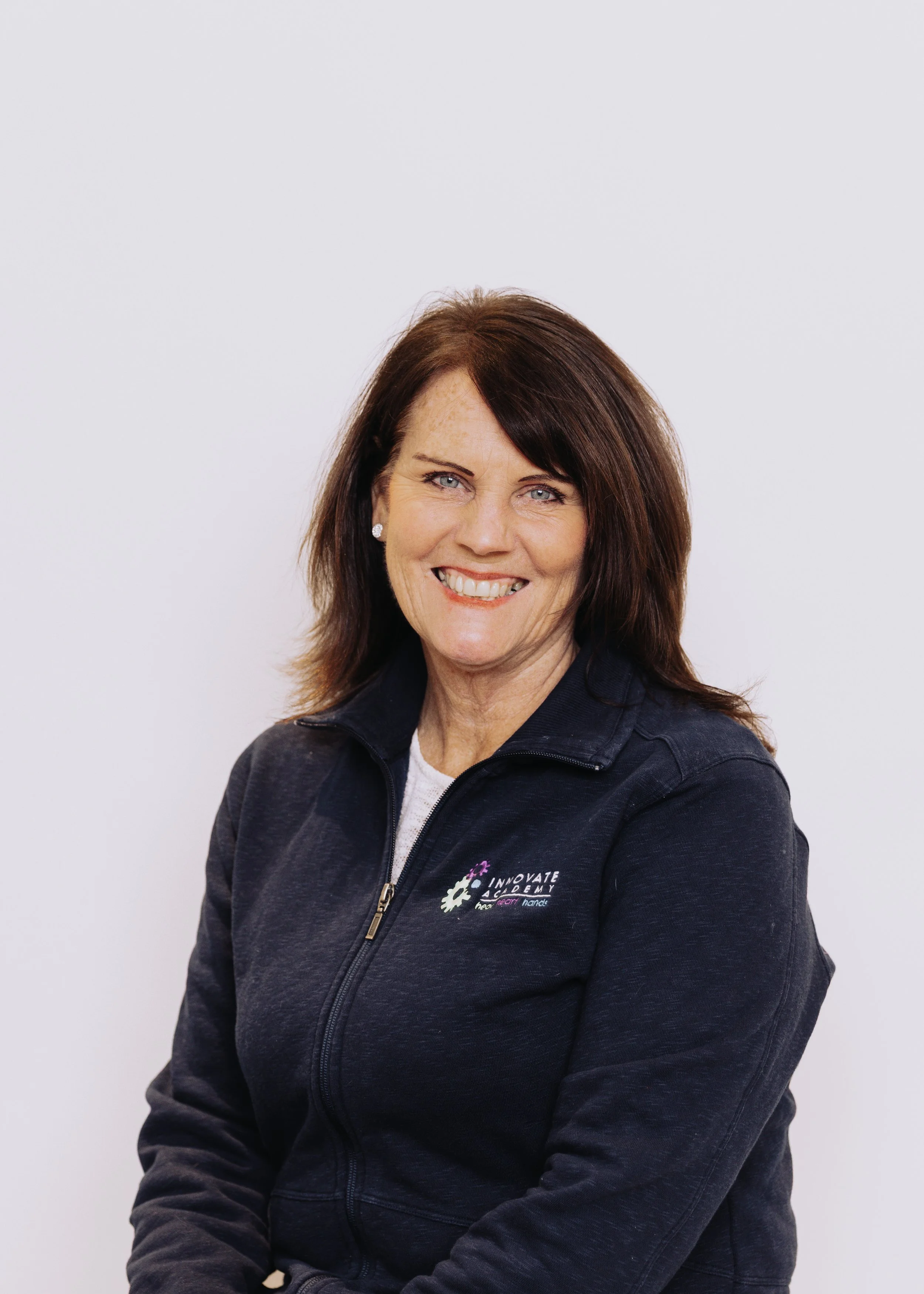 Portrait of a smiling woman with shoulder-length brown hair, wearing a navy blue zip-up jacket with a logo on the chest, against a plain white background.
