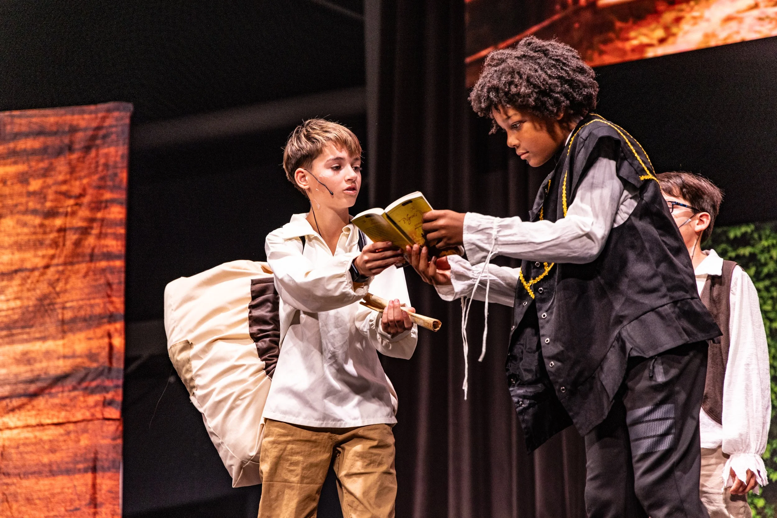Children performing on stage: boy with backpack and microphone headset holding a stick, boy with afro wig in a black costume reading from a book, another boy with glasses in the background.