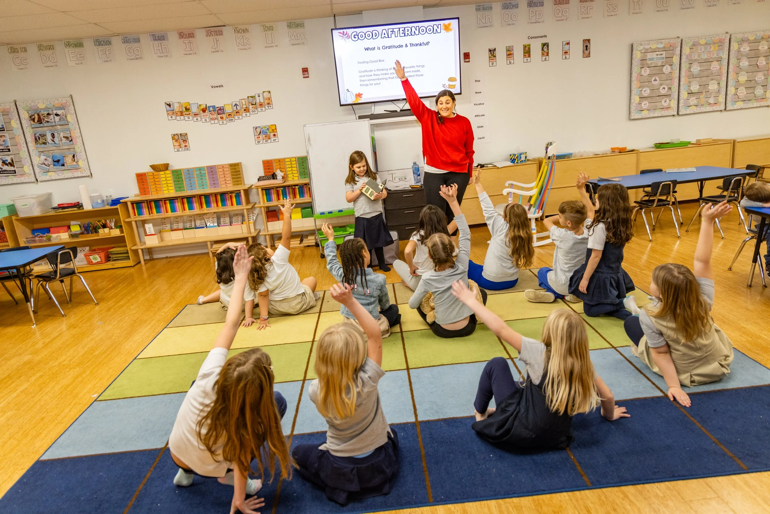 A classroom with children sitting on a colorful carpet, raising their hands, as a teacher in a red sweater stands at the front, smiling. The teacher points to a large screen displaying a message about gratitude and thankfulness, with a young girl holding a book nearby. The classroom has educational posters, shelves with books and supplies, and is brightly lit.