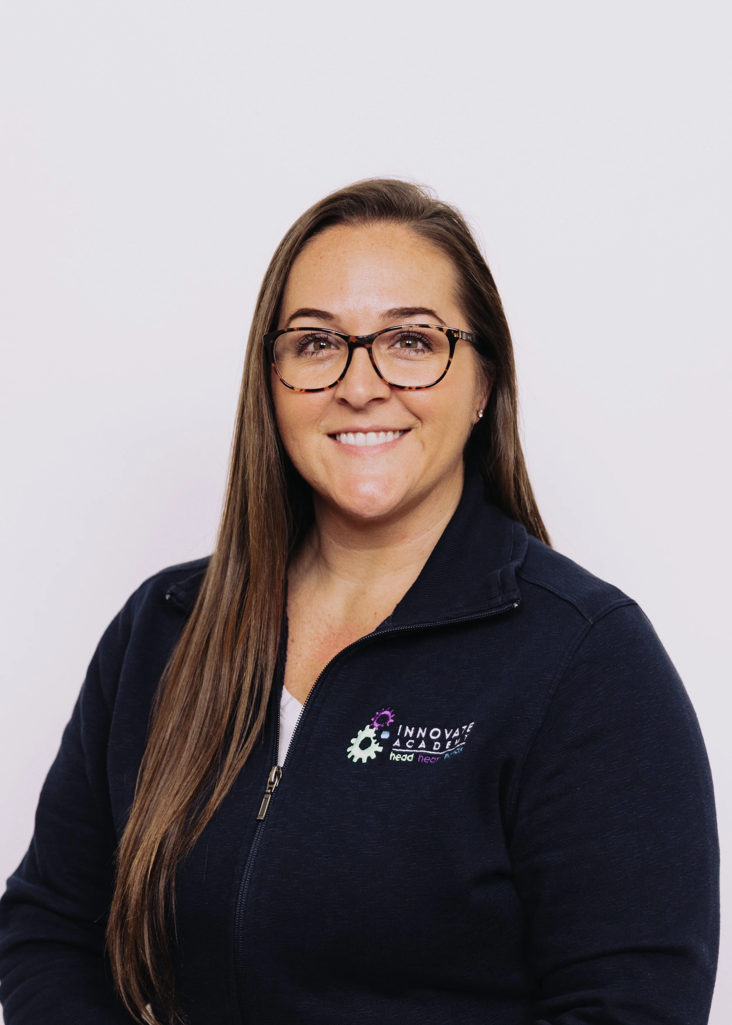A woman with long brown hair, glasses, and a navy fleece jacket with a logo, smiling against a white background.