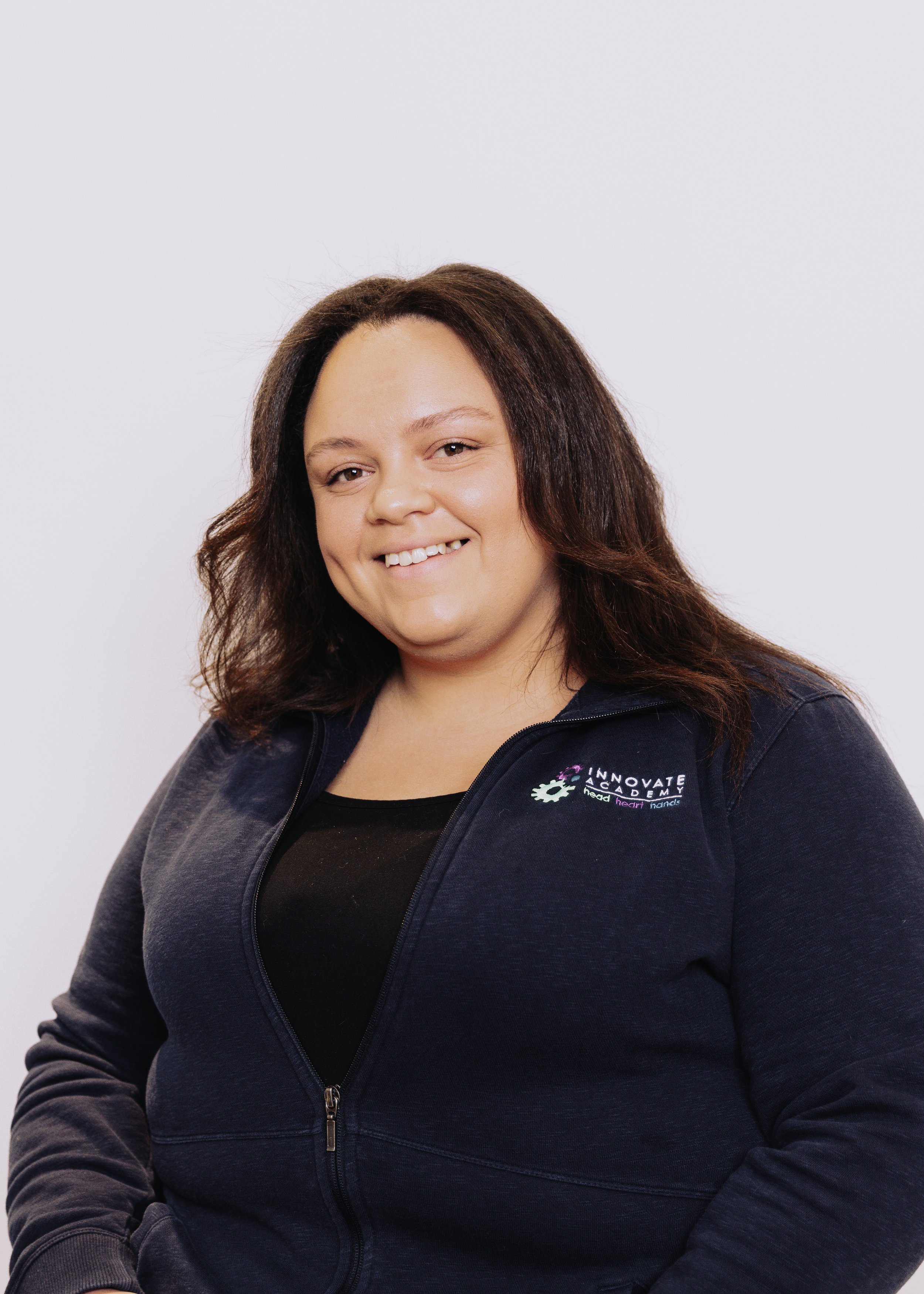 A woman with shoulder-length brown hair smiling at the camera, wearing a navy blue zip-up jacket with an 'Innovate Academy' logo on the left chest and a black shirt underneath, standing against a plain white background.