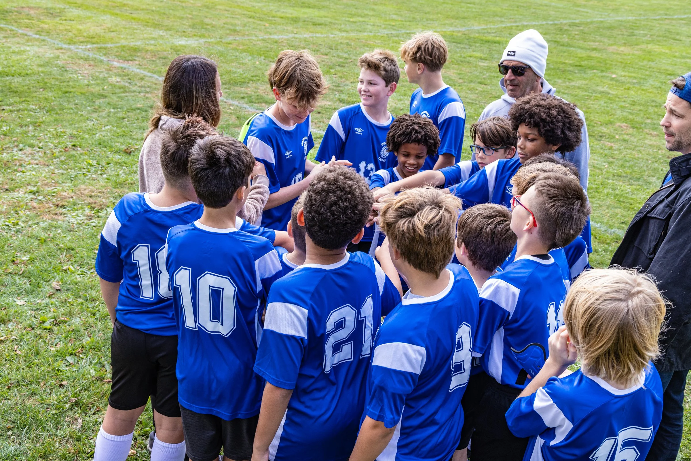Youth soccer team in blue uniforms gathered in a circle on the field, receiving a pep talk from their coach before the game.