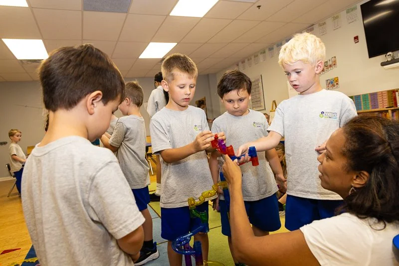 Group of young children gathered around a woman playing with a colorful marble run in a classroom.