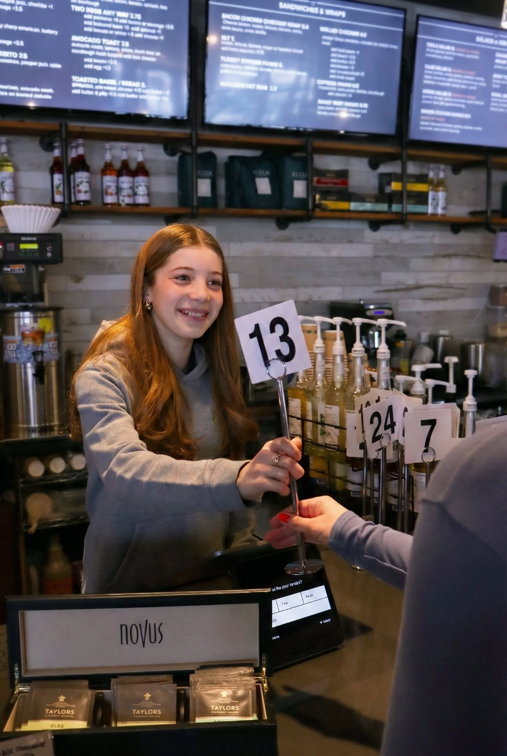 A smiling woman completes a transaction at a cafe counter, exchanging a number card '13' with a customer. Behind her are shelves with bottles of syrup and a digital menu screen overhead.