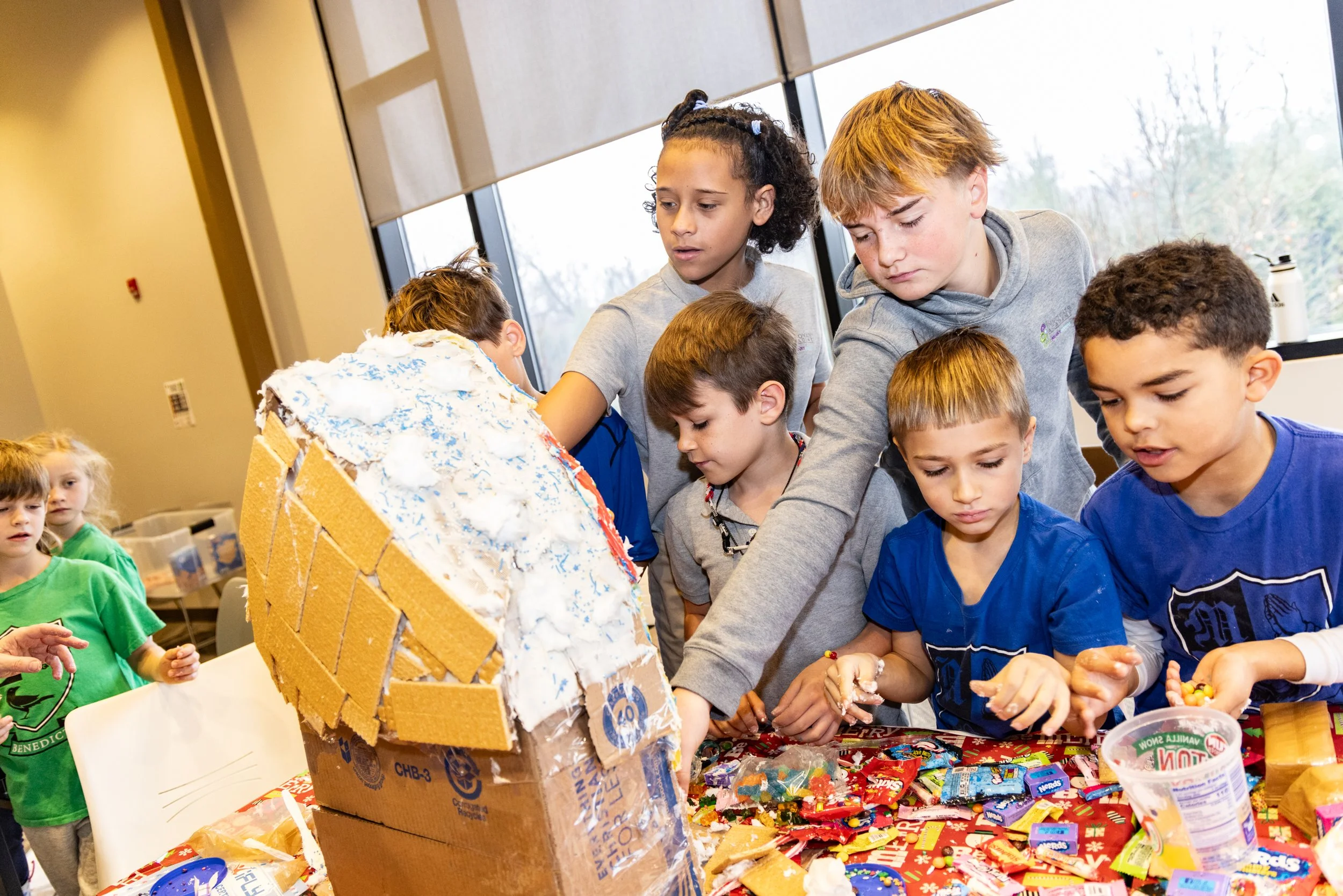 Children and teenagers gathered around a table, building or decorating a large gingerbread house with candies and icing during a festive event.