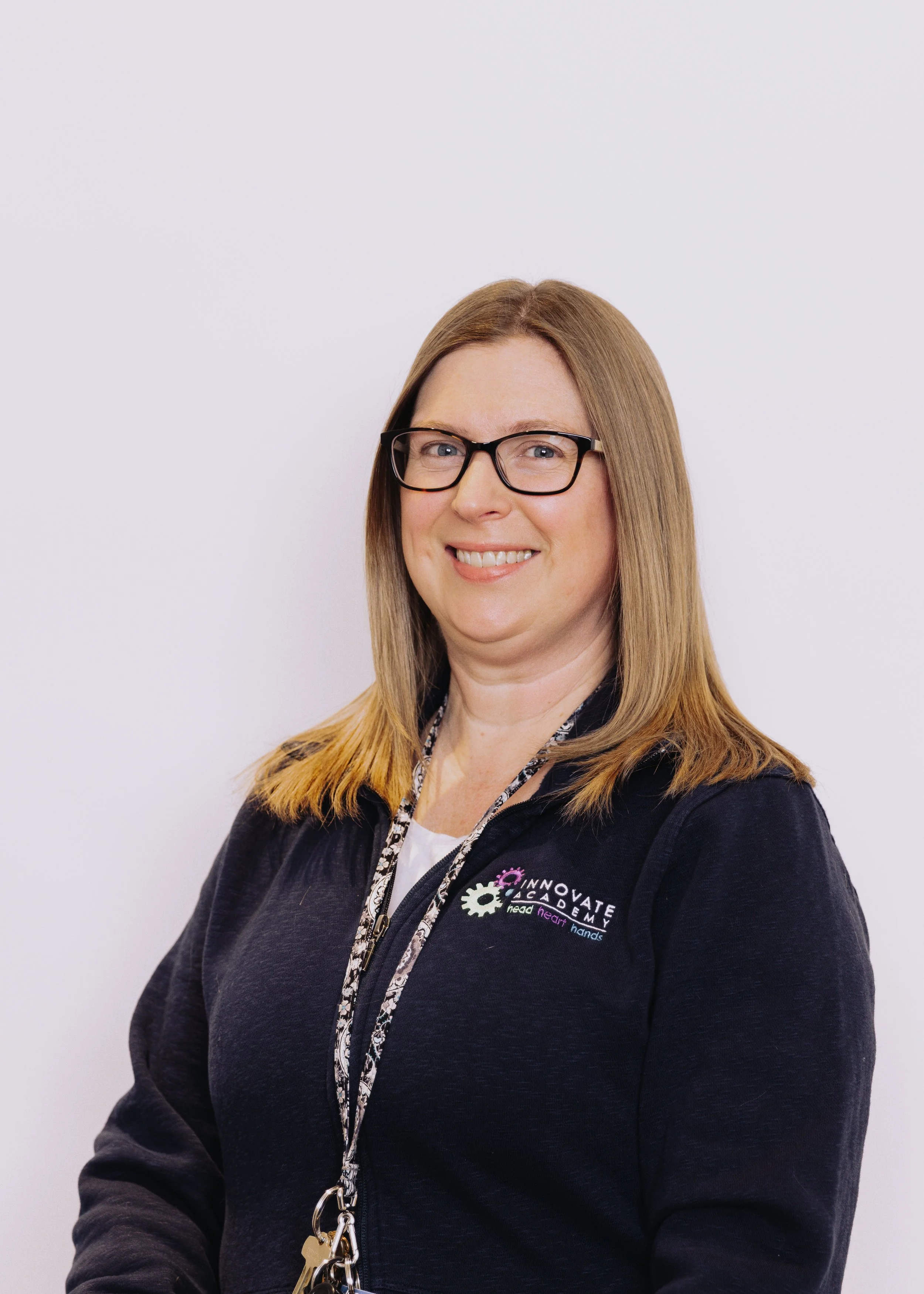 A woman with shoulder-length blonde hair, wearing glasses, a black jacket with a logo that says 'Innovate Academy' and a lanyard with keys, smiling in front of a white background.