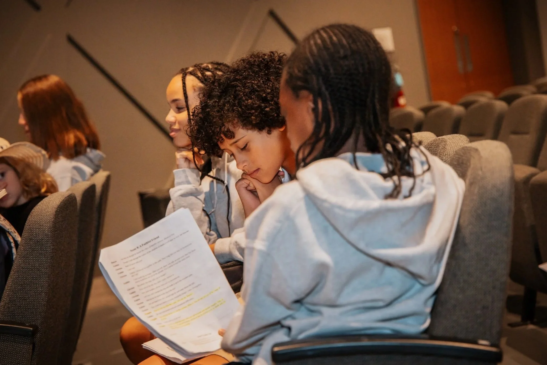 Kids sitting in auditorium seats, reading and listening to something.