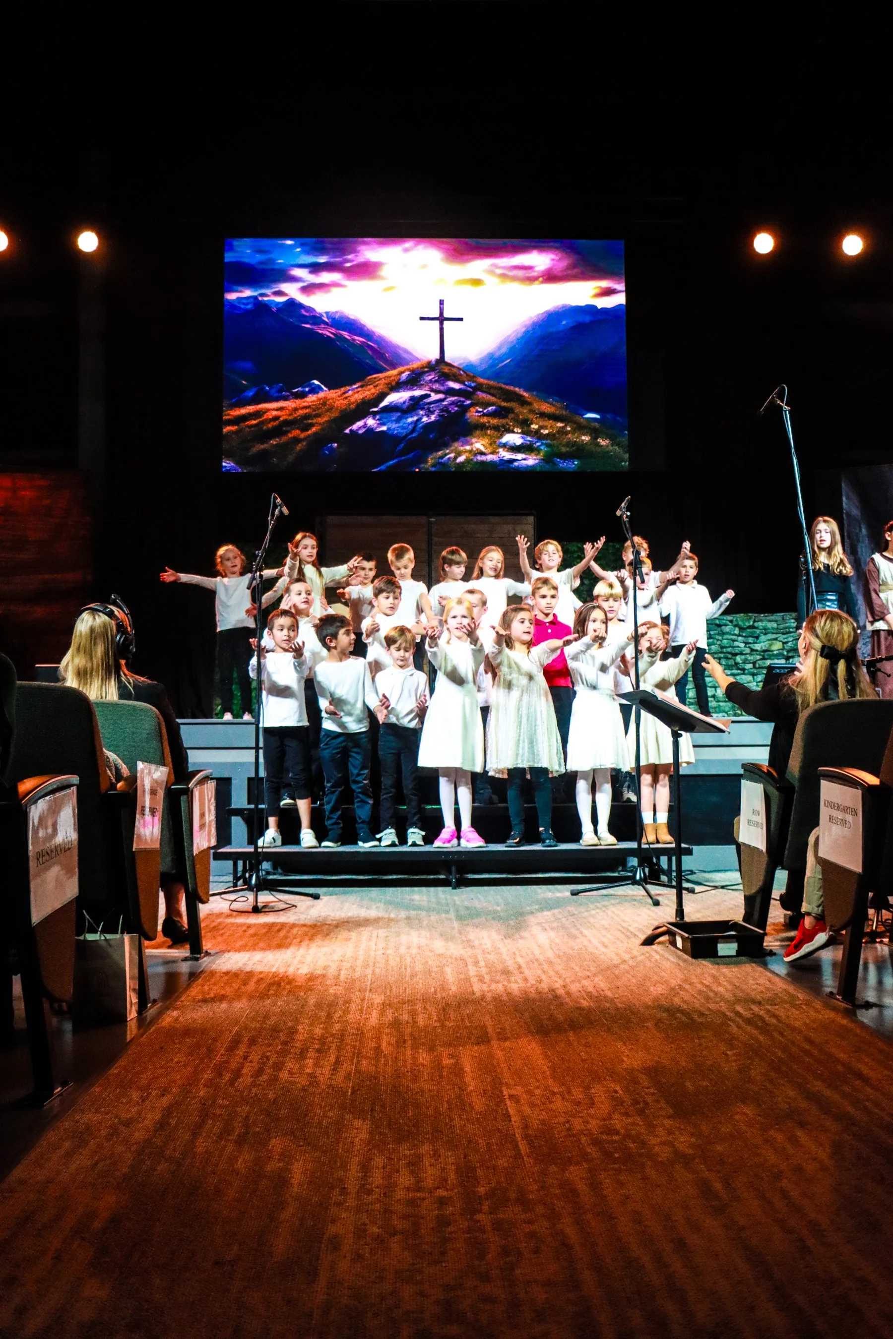 A group of children performing on stage in a church or school setting with a large screen in the background displaying a cross on a hill at sunset.