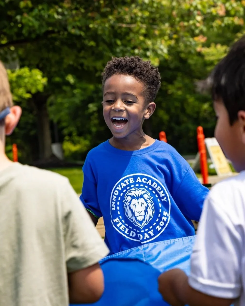 Smiling young boy in a blue shirt with a lion logo, standing outdoors with two other children, on a sunny day surrounded by green trees.