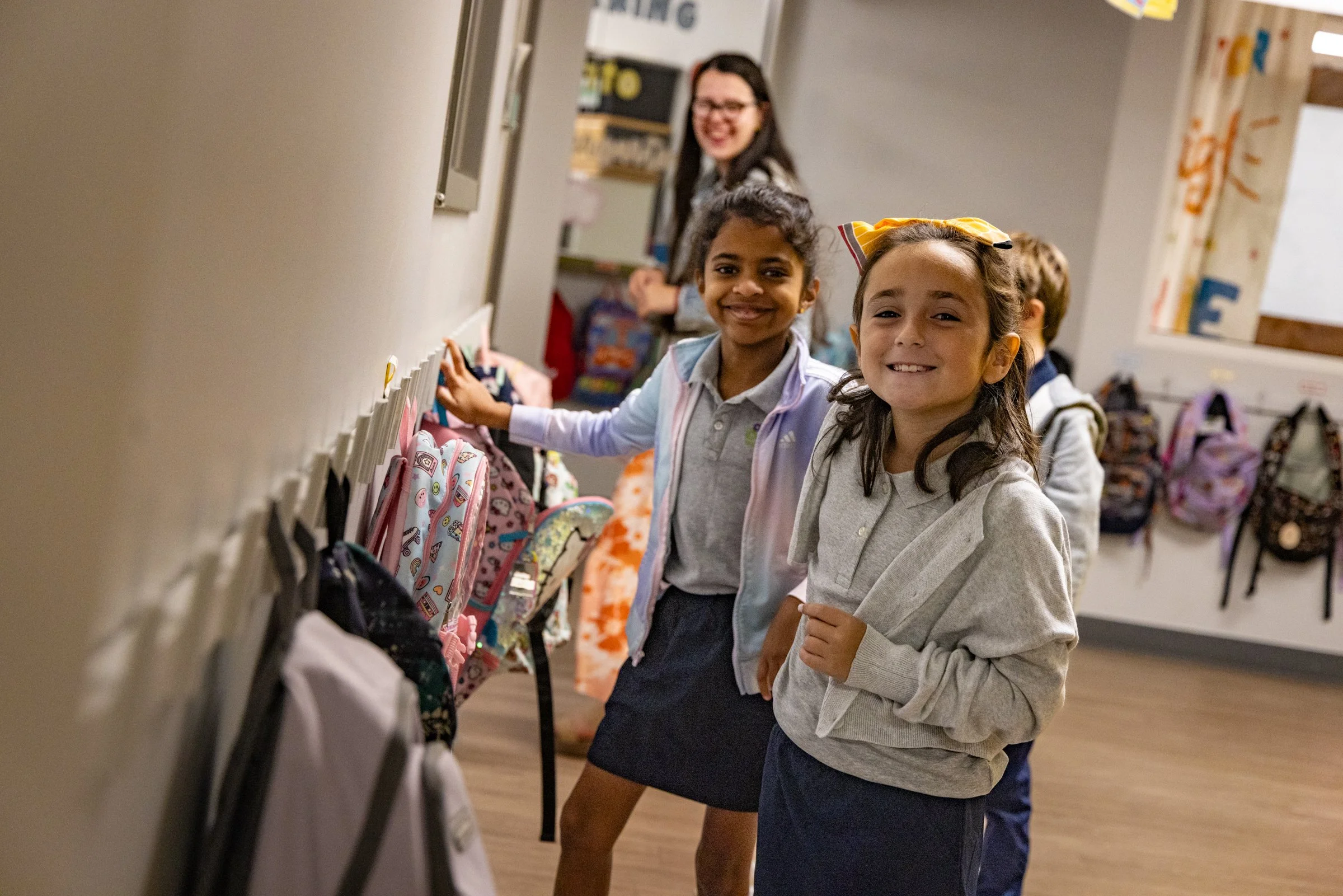 Two smiling young girls with backpacks browsing backpacks on hooks in a school hallway, with a teacher in the background.
