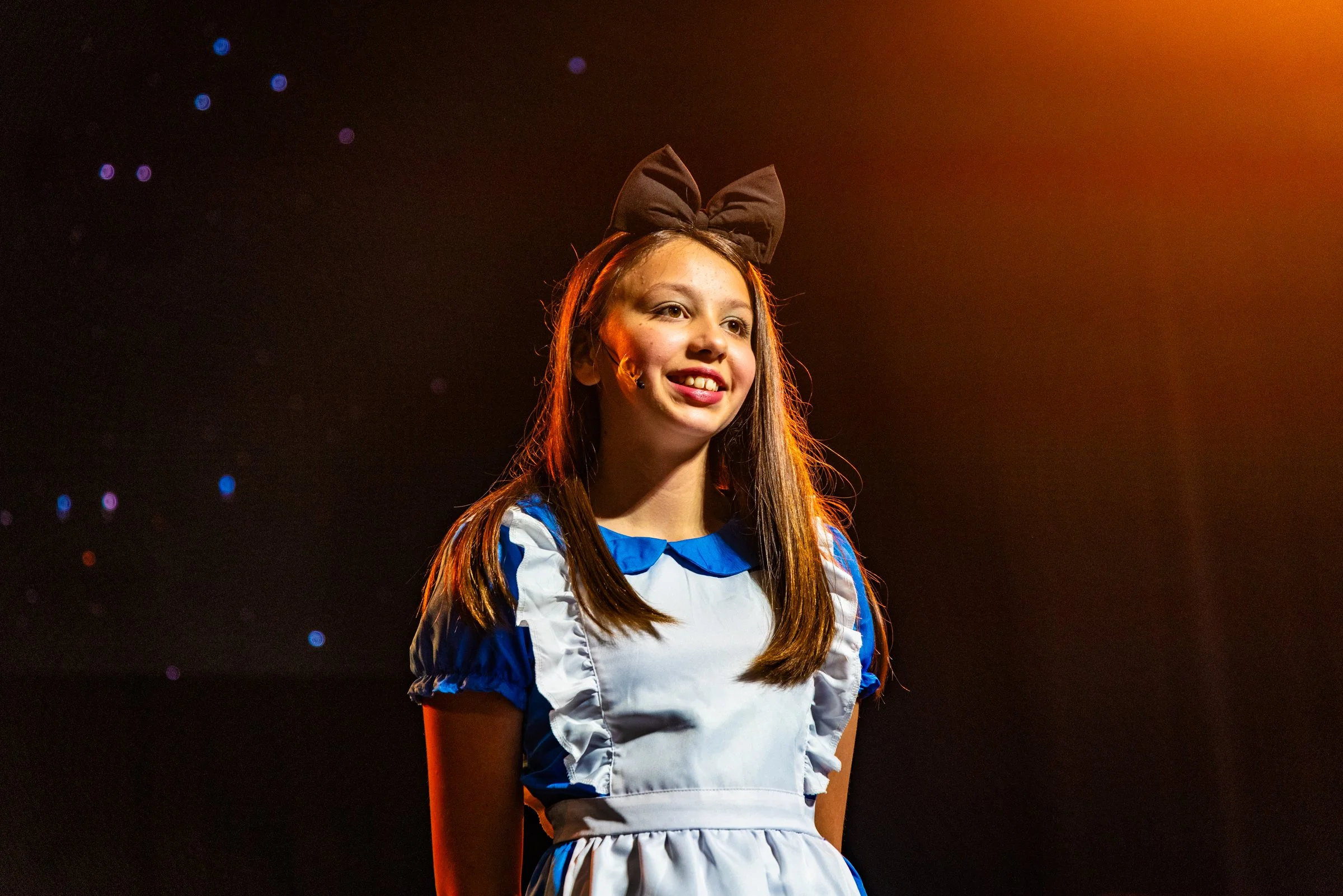 Young girl dressed as Dorothy from The Wizard of Oz, smiling on stage with a black bow in her hair, under warm stage lighting.
