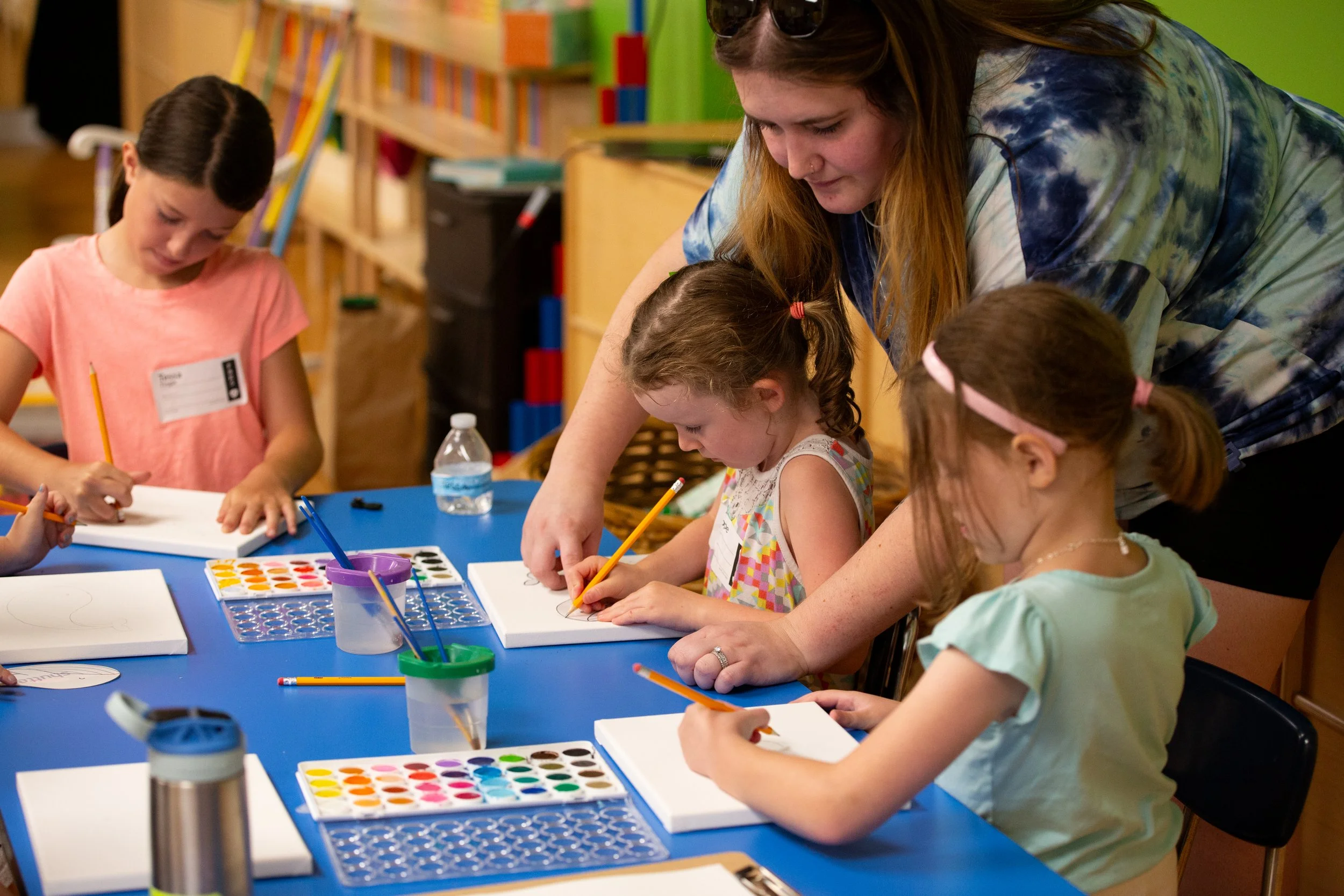 A group of young girls and two adults drawing and painting at a blue table covered with art supplies like watercolors, paintbrushes, and paper in a colorful classroom.