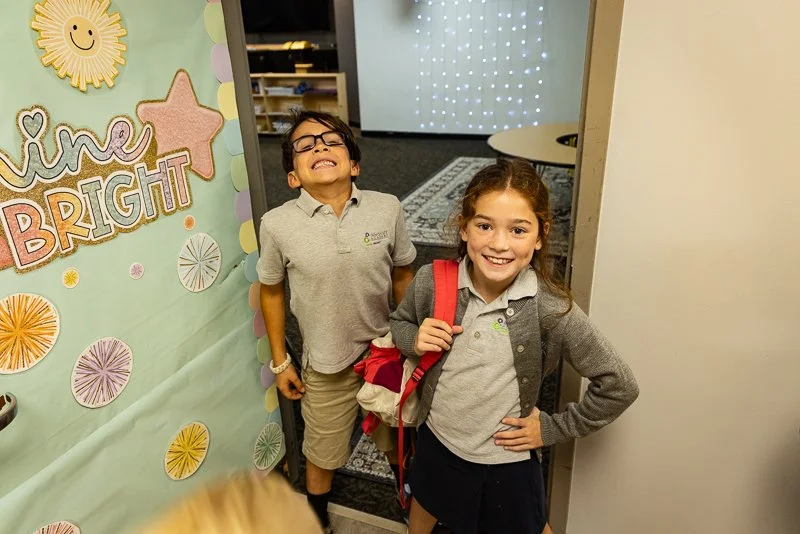Two children in school uniforms smiling and standing at the entrance of a classroom or hallway.