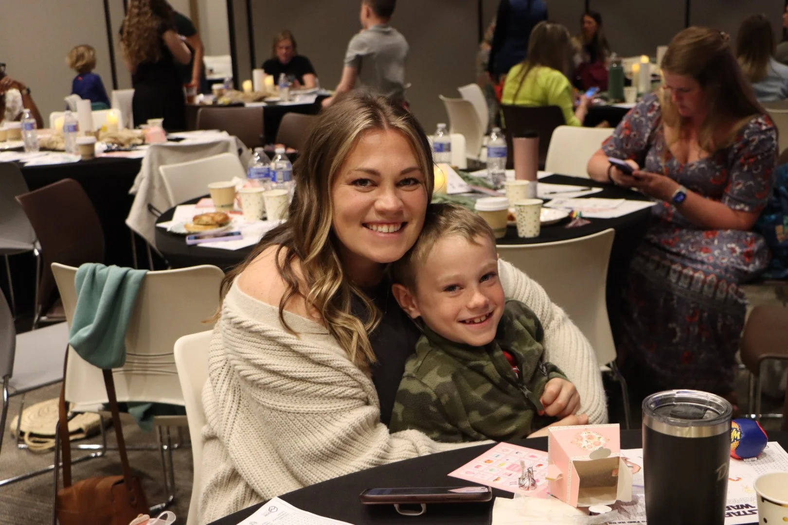 A woman and young boy smiling and sitting close together at a table during a gathering, with other people in the background