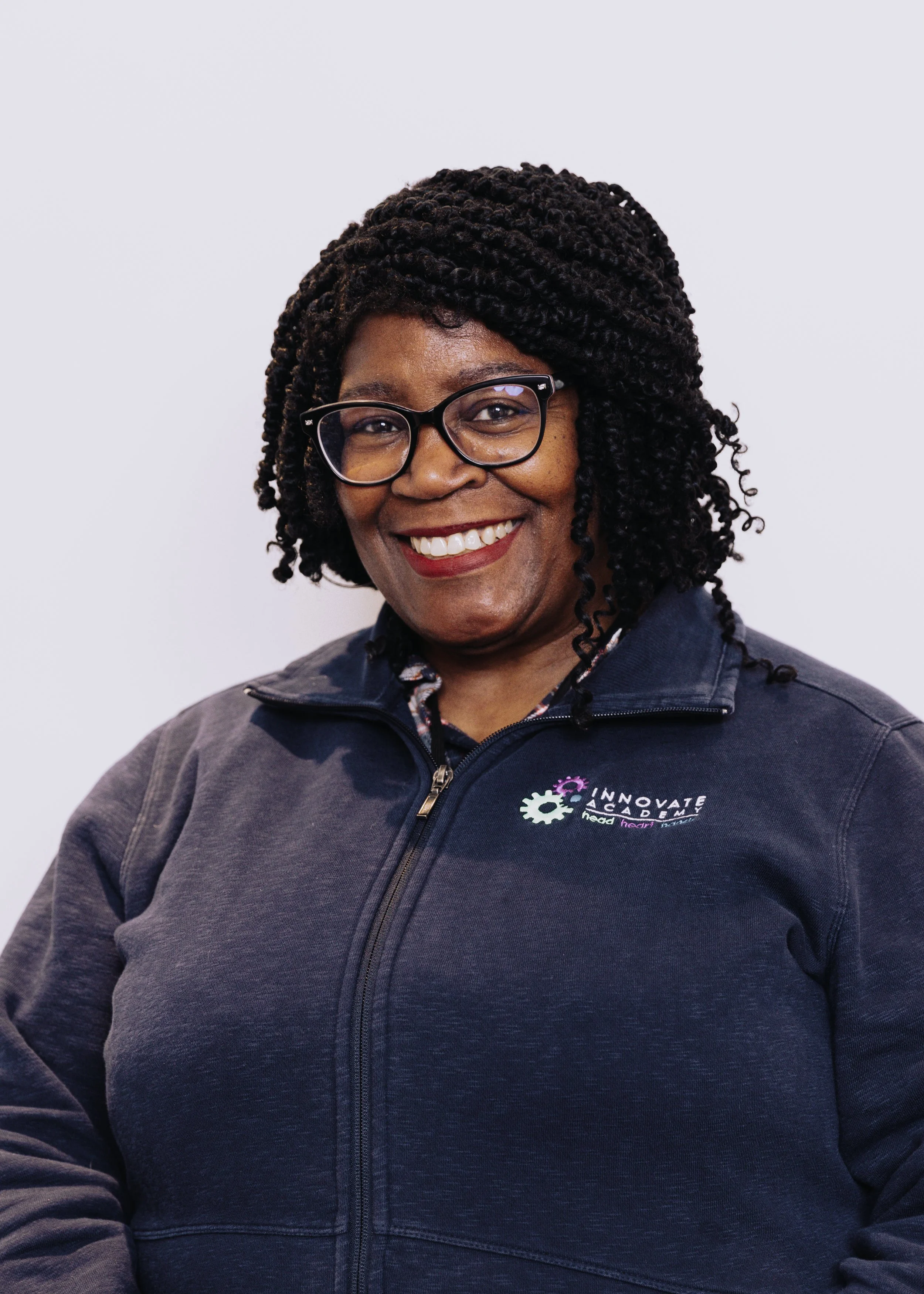 A smiling African American woman wearing glasses and a dark blue zip-up jacket with a logo on the chest, standing against a plain white background.
