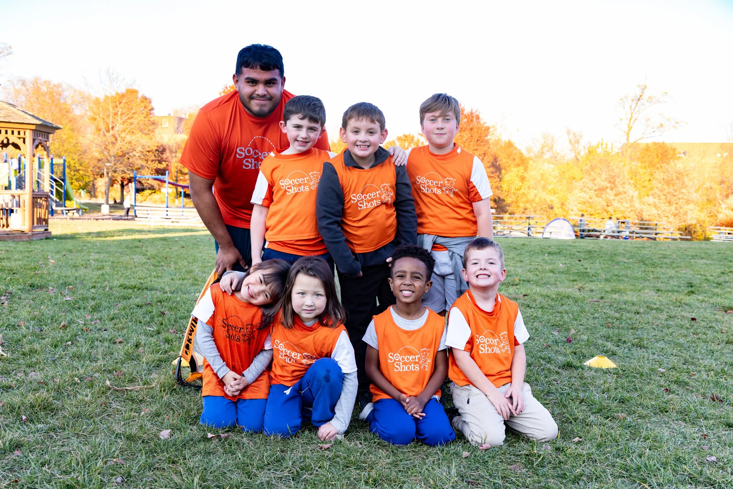 A coach and a group of young children with matching orange soccer jerseys posing outdoors on a grassy field during autumn.