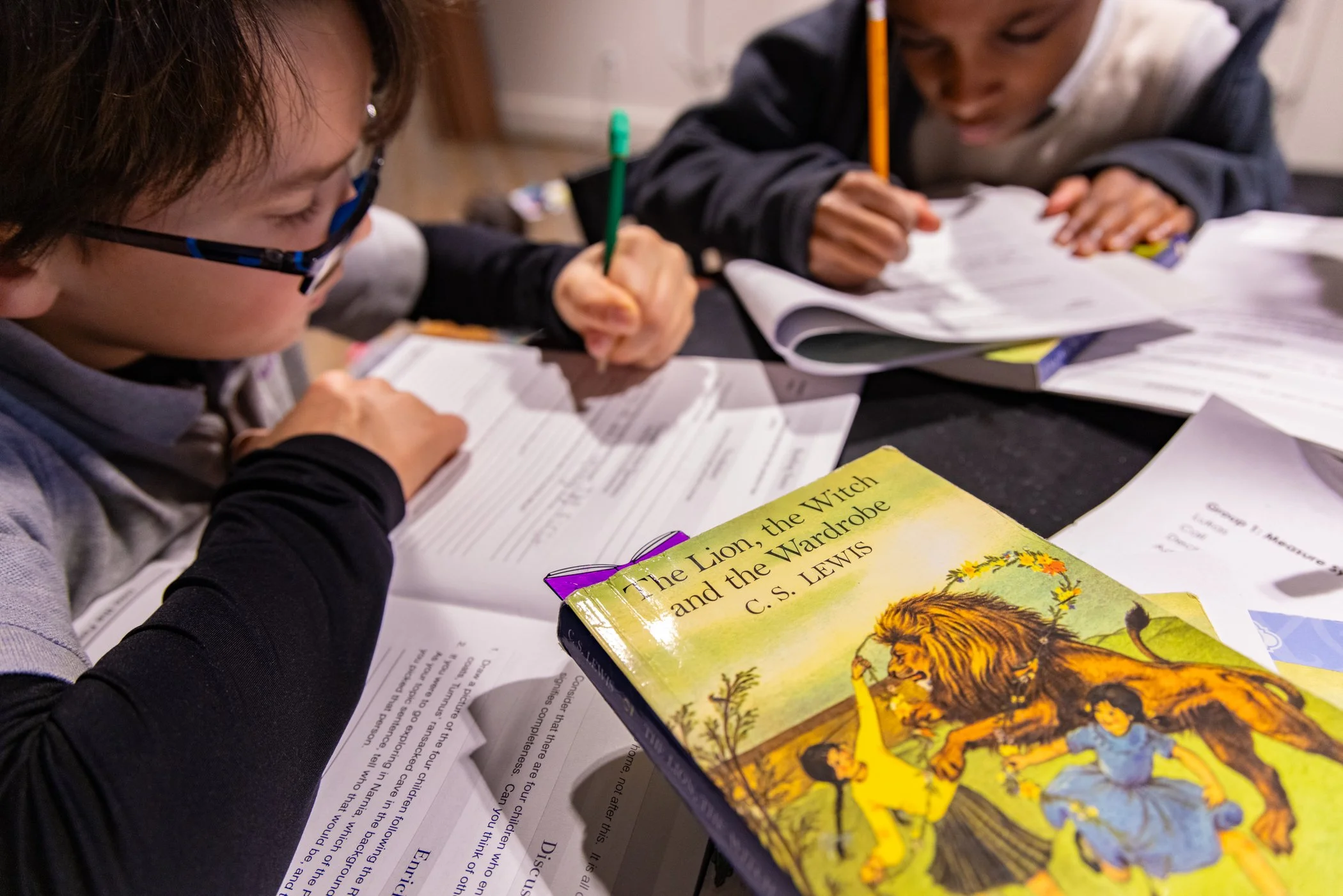 Two children sitting at a table with open books and papers, one child wearing glasses, studying and working on assignments, with a colorful book titled 'The Lion, the Witch and the Wardrobe' in the foreground.