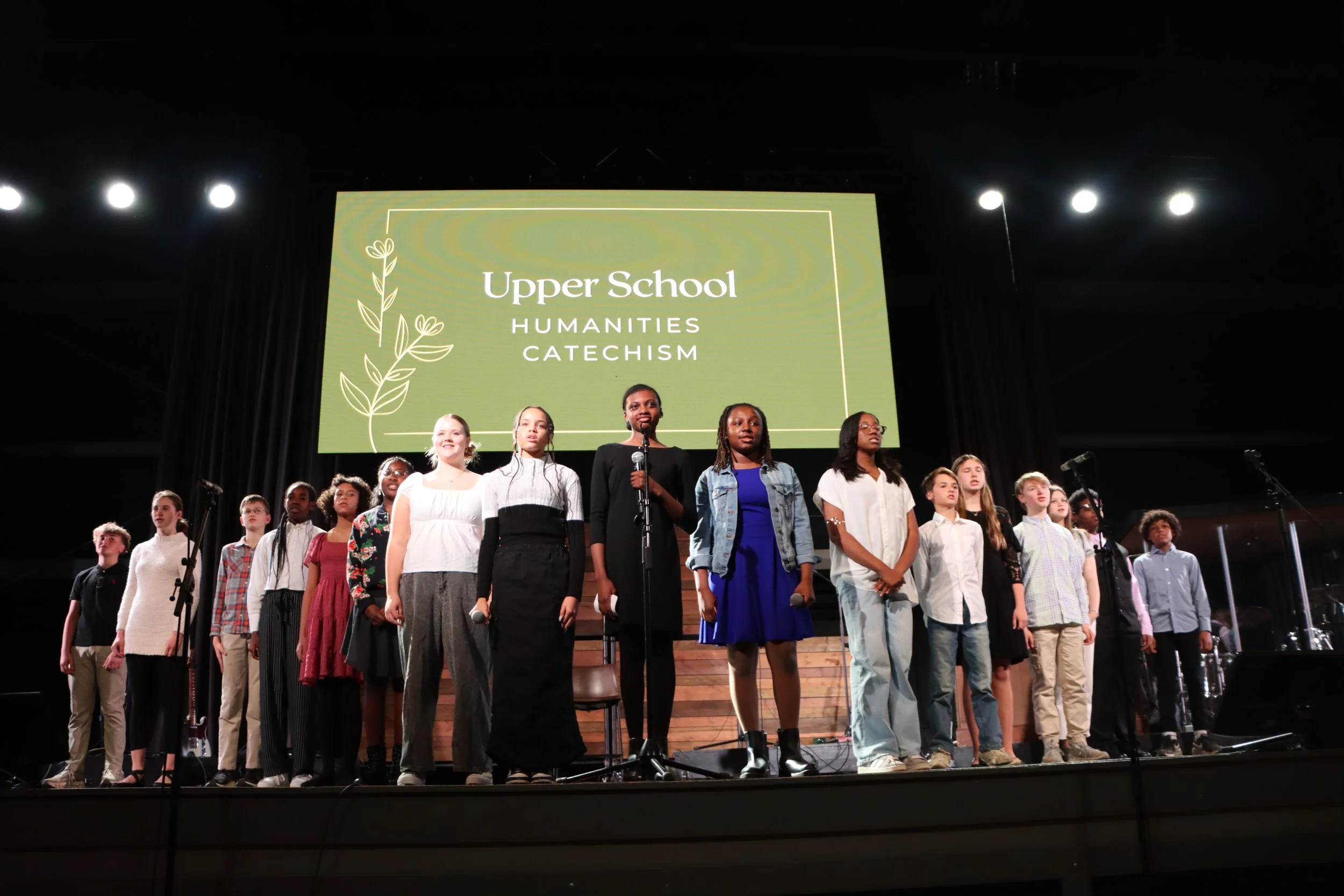A group of students standing on stage during a school event, with a large screen behind them displaying 'Upper School Humanities Catechism' and a decorative plant illustration.