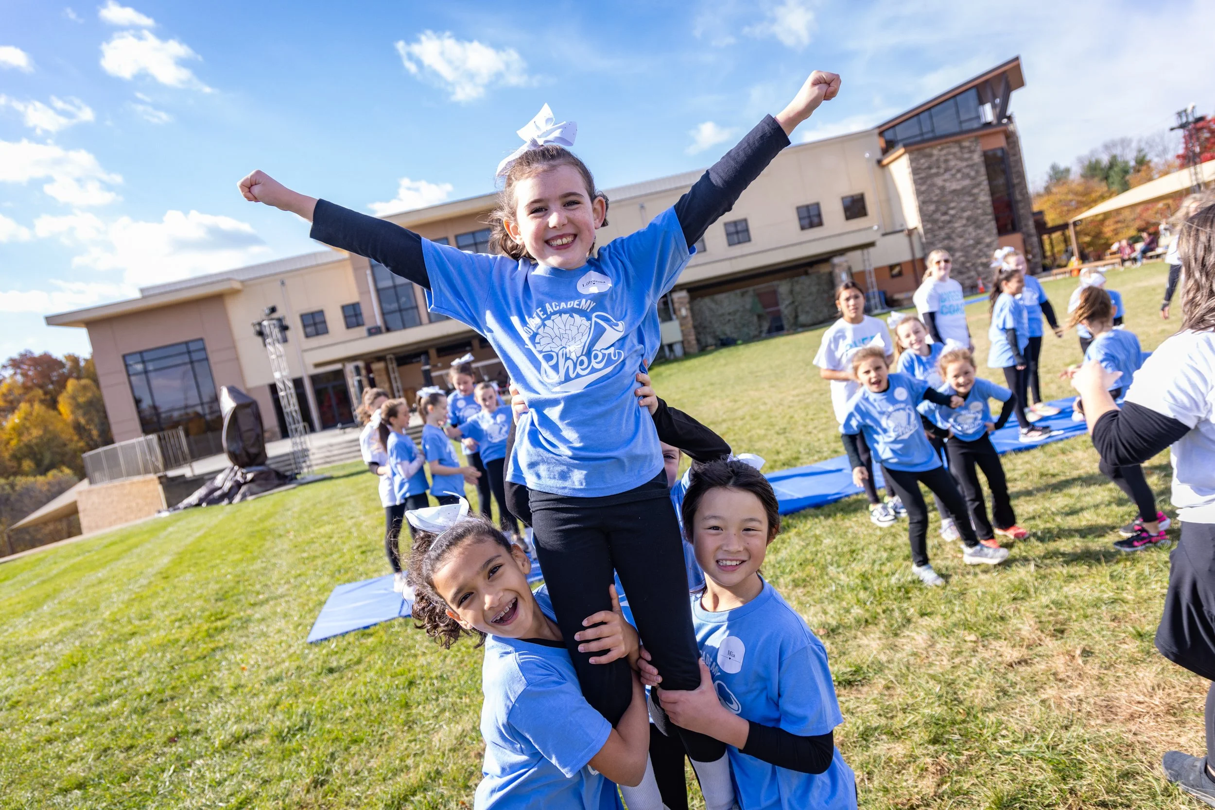 Cheerleaders perform stunts outdoors on a grassy field with a school building in the background. The girls wear blue cheer uniforms and the weather is sunny with a blue sky.