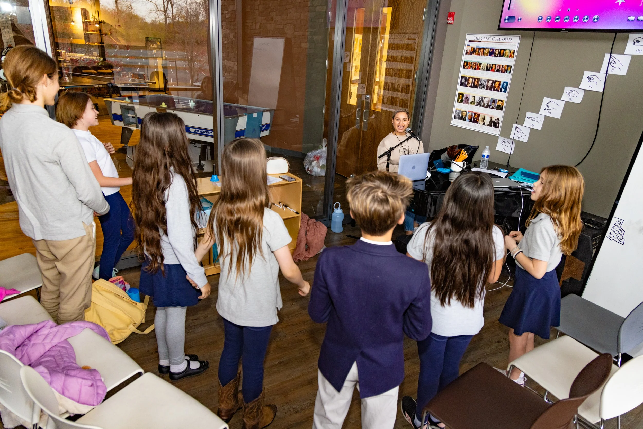 Children gathered around a woman at a piano during a music lesson, with sheet music and educational posters on the wall in a classroom setting.