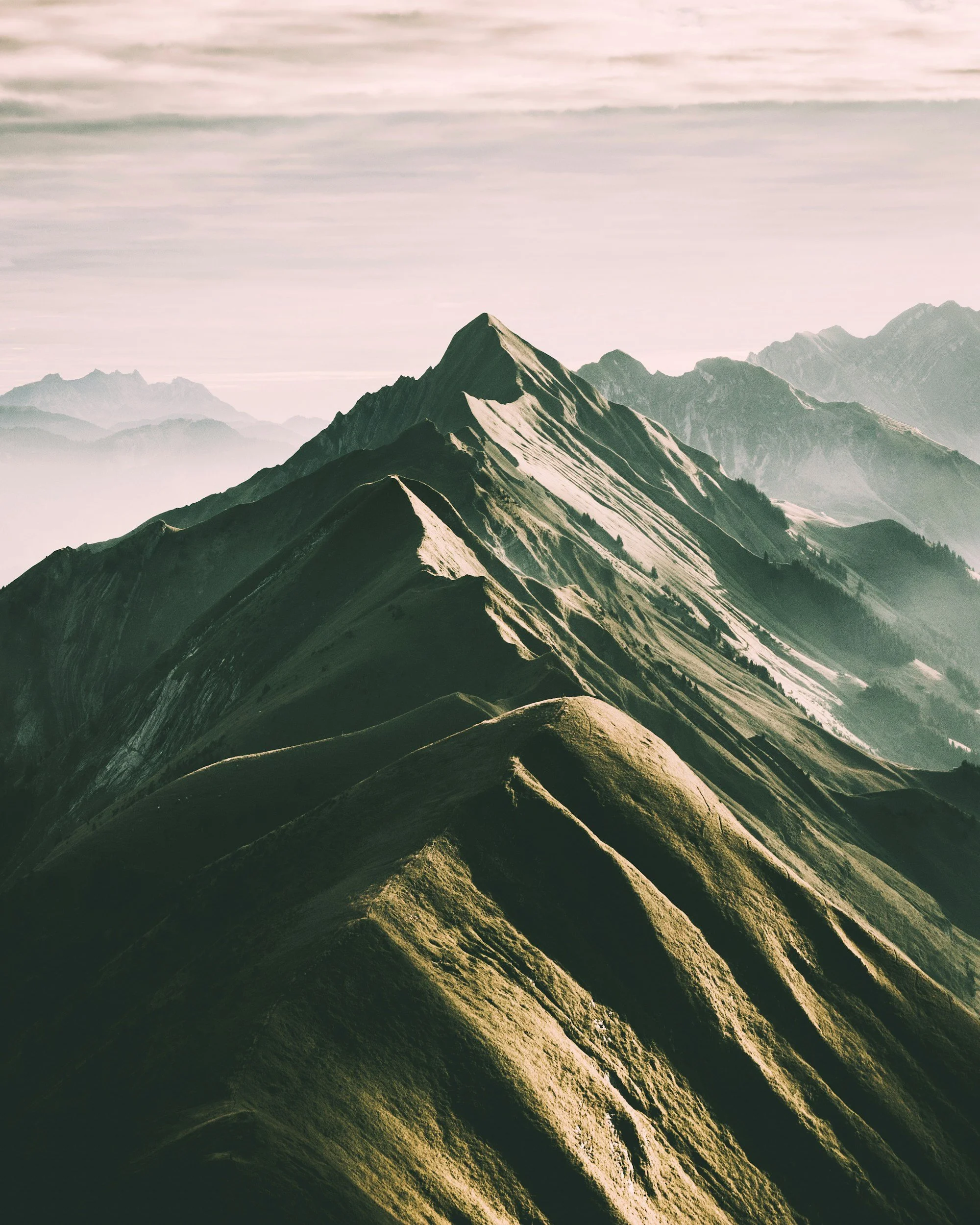 A panoramic view of mountain peaks with green slopes and some snow, with layered mountain ranges in the background under a slightly cloudy sky.