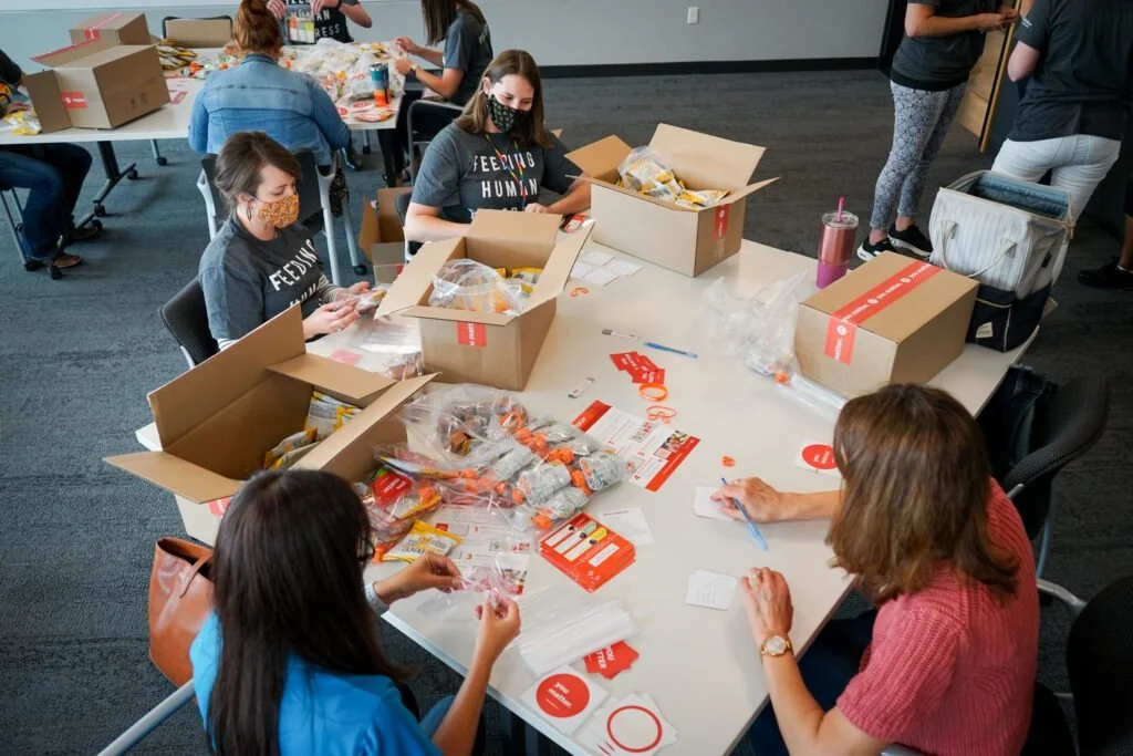 Group of women and girls assembling safety kits or packages at a large table in an indoor setting.
