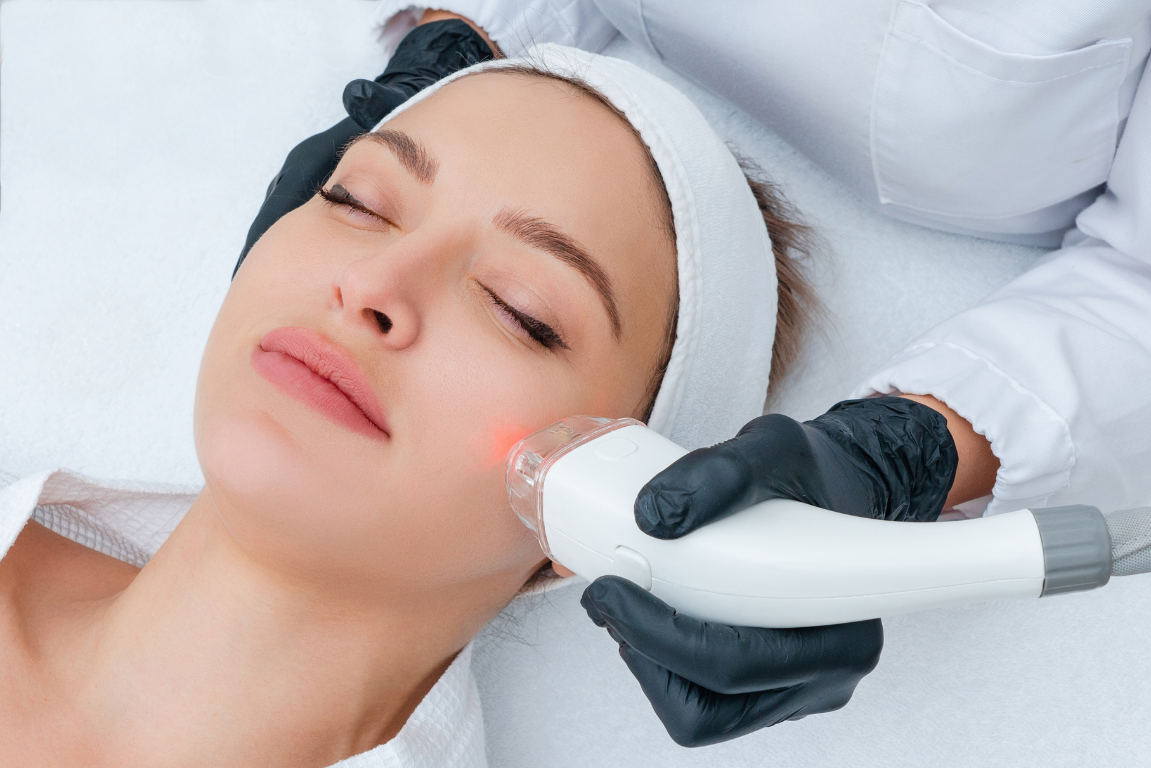 A woman receiving a facial skin treatment with a handheld device emitting red light, lying peacefully with eyes closed, in a spa or clinic setting.