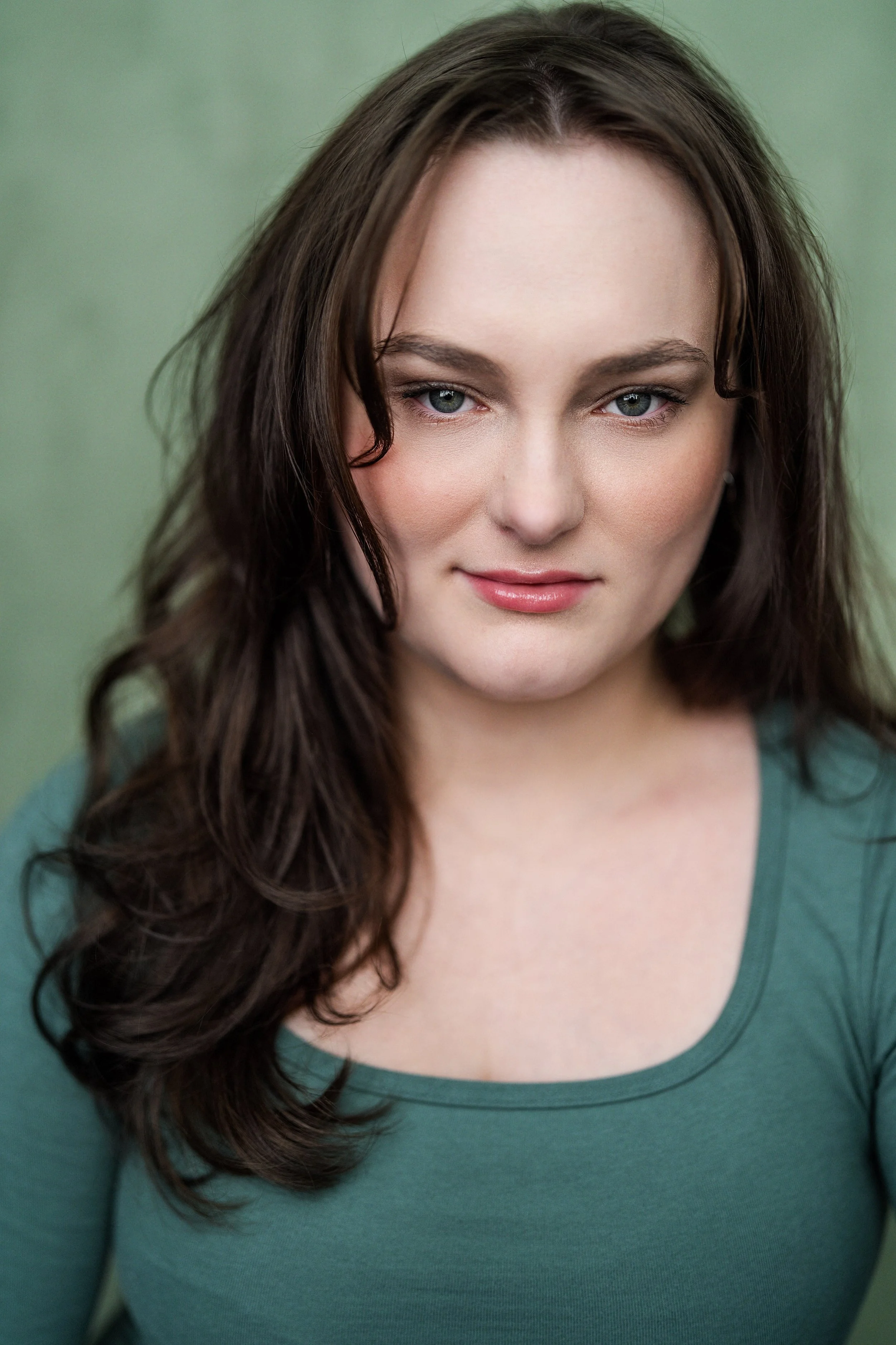 A young woman with long wavy brown hair and blue eyes, wearing a green top, smiling softly against a green background.