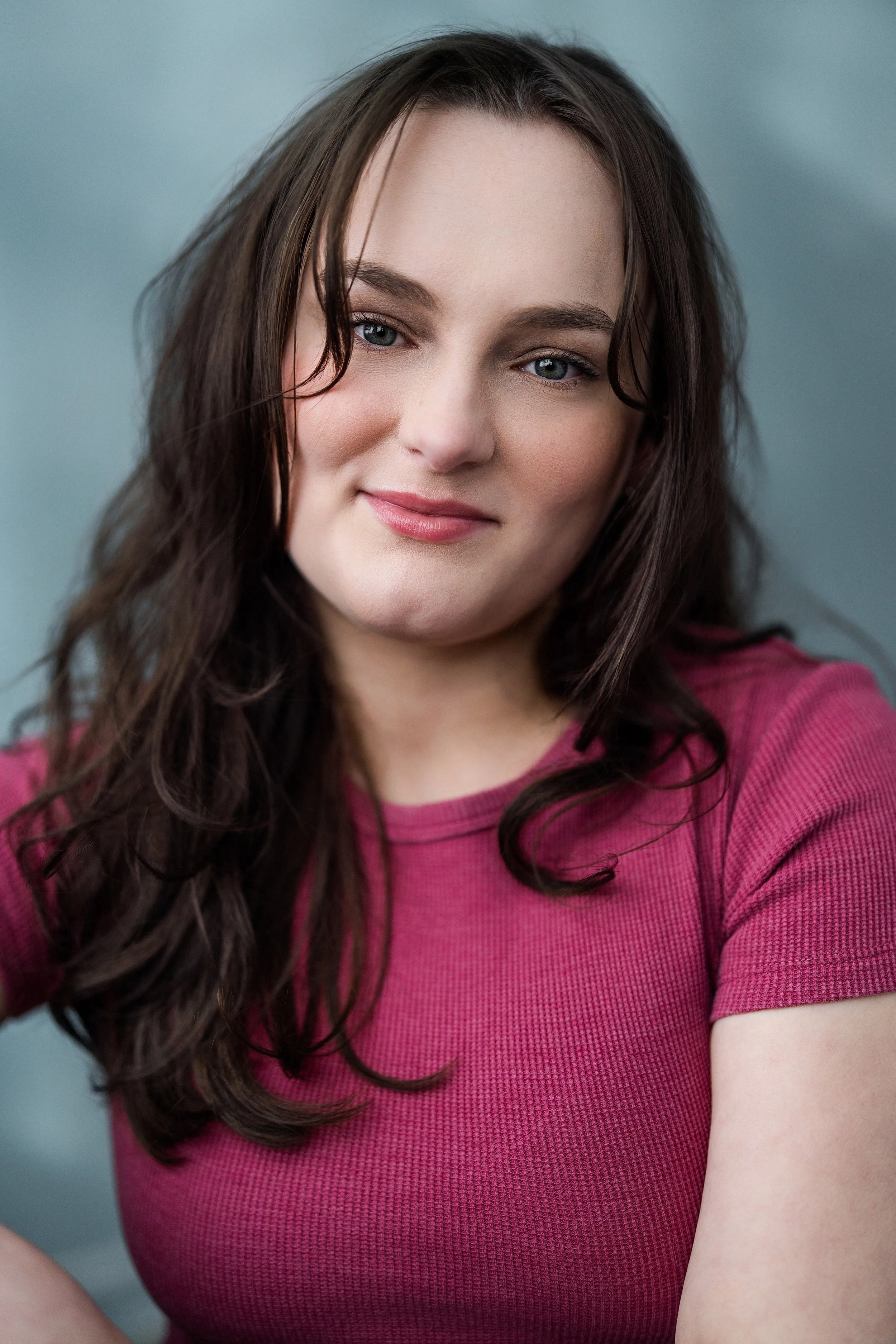 A young woman with brown hair and blue eyes wearing a pink shirt, smiling softly at the camera.
