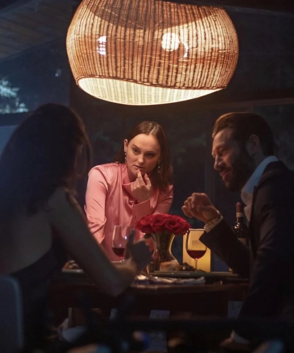 Three people seated around a dinner table with red wine, a vase of red roses, and a plate of food at night, illuminated by a large hanging wicker light.
