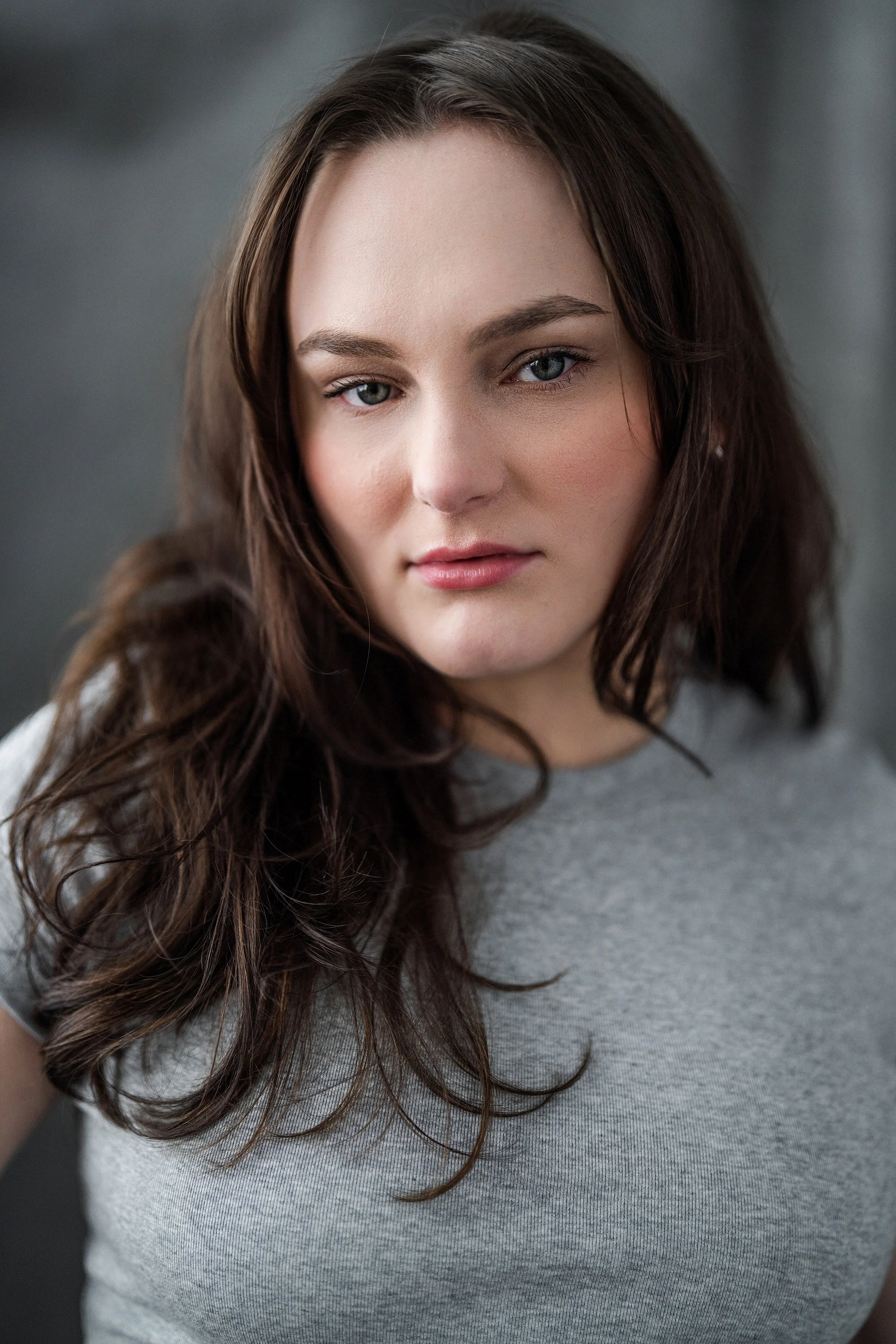 Close-up portrait of a young woman with long, wavy brown hair wearing a gray shirt, looking directly at the camera against a blurred dark background.