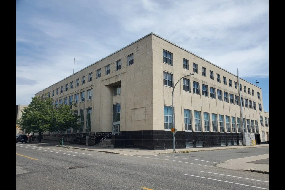 A beige multi-story building with numerous windows, a black lower section, and a small staircase leading to the entrance, located on a corner lot with streetlights and trees nearby.