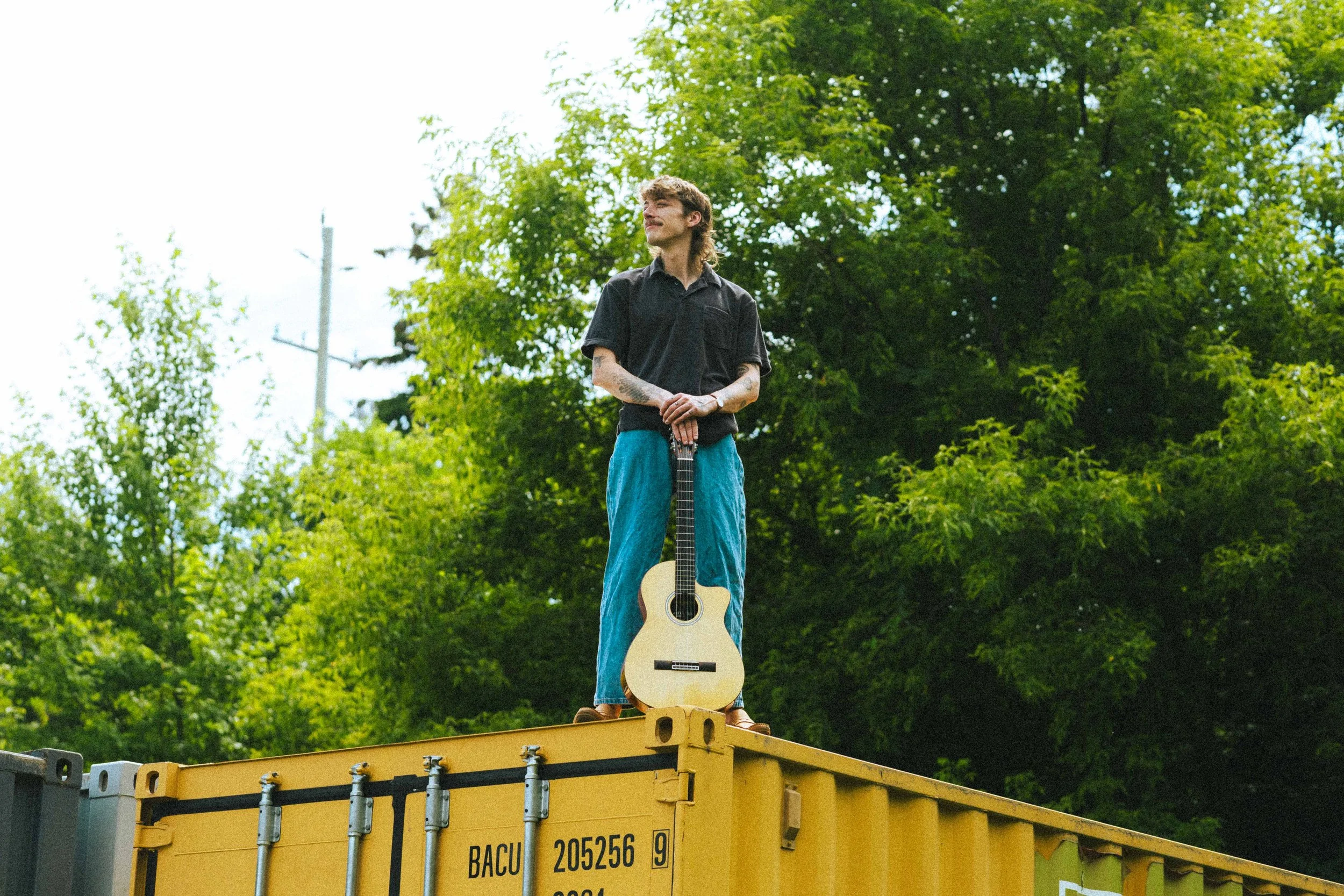 A man standing on top of a yellow shipping container, holding an acoustic guitar, surrounded by green trees.