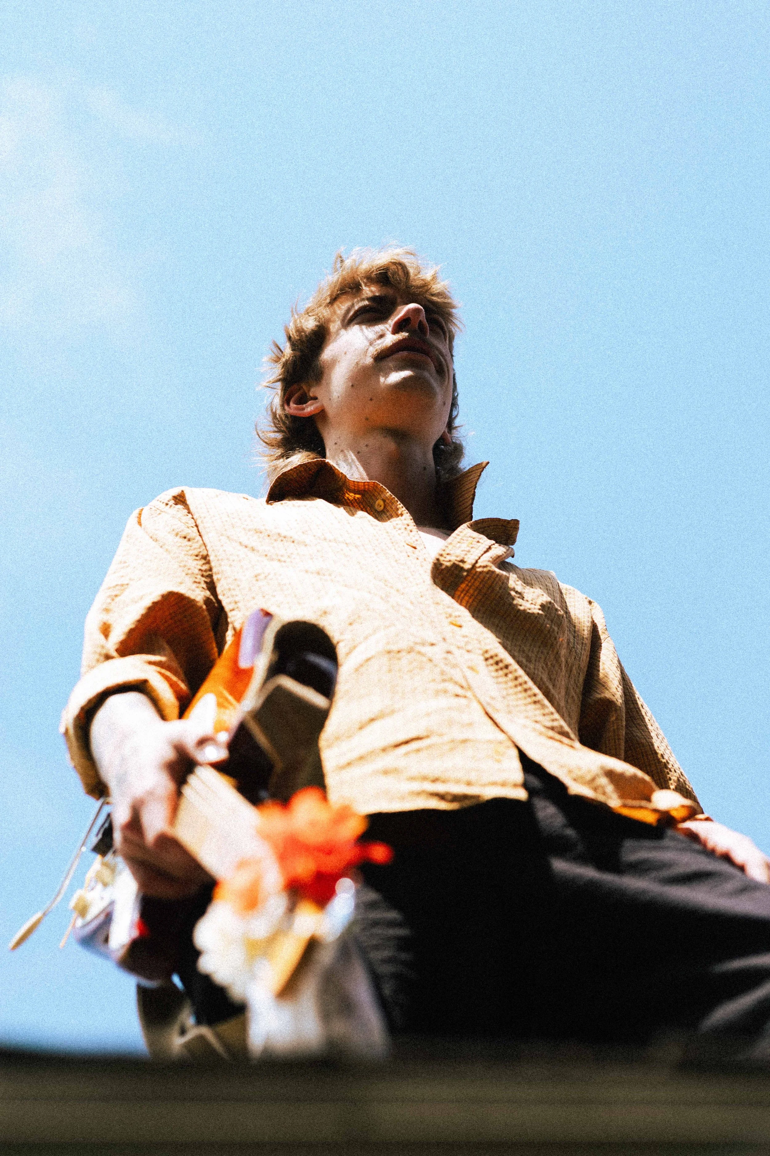 Low-angle view of a young man looking up, wearing a beige jacket, against a clear blue sky.