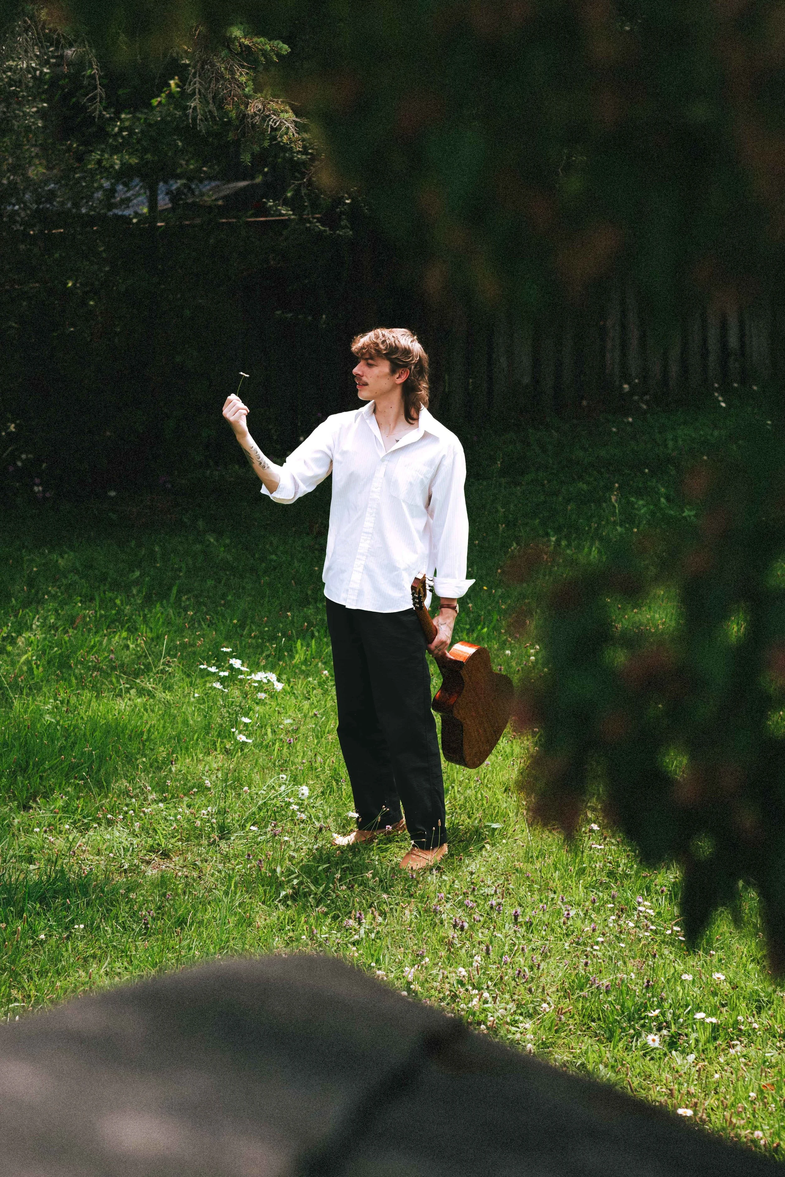 A young man with long hair, a white shirt, and black pants standing on grass in a park or garden, holding a small item up in one hand and a guitar in the other.