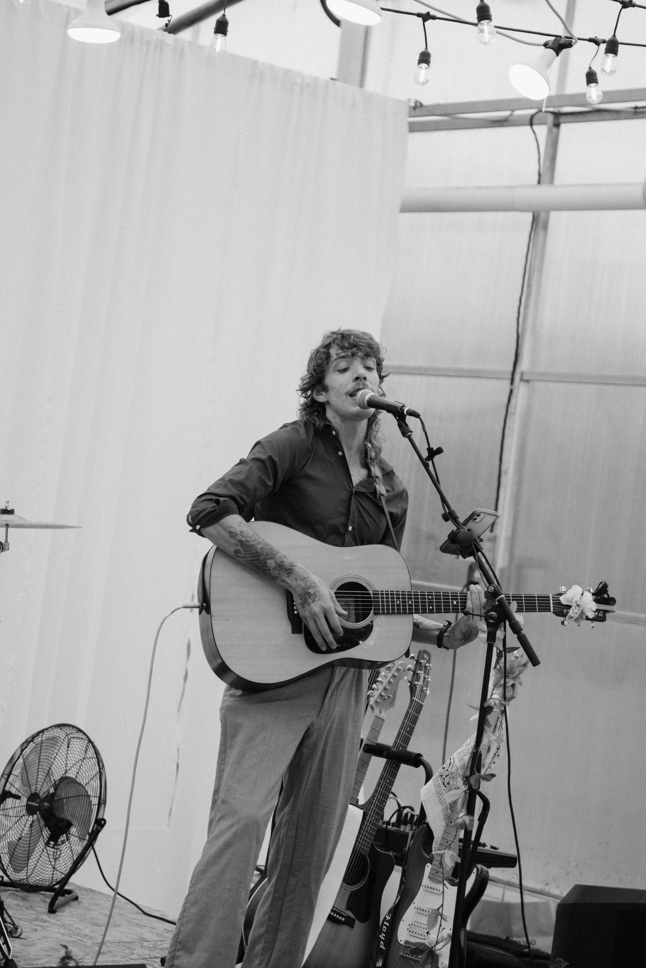 A musician with wavy hair and tattoos is singing and playing an acoustic guitar on stage, with a microphone. A fan and two other guitars are visible in the background, with string lights hanging overhead.